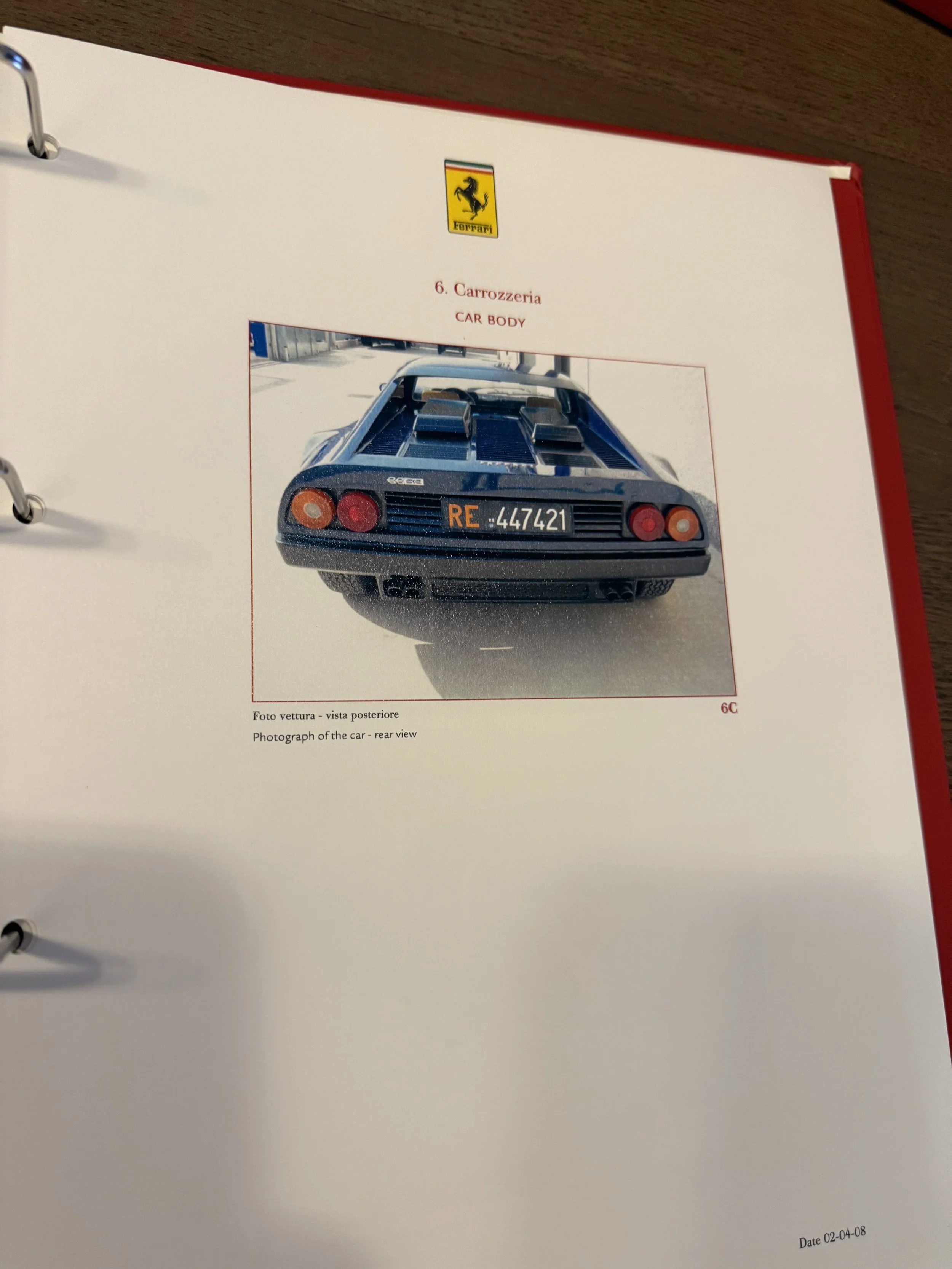 Photograph of a classic black Ferrari car seen from the rear, with round taillights and a license plate that reads "RE 447421." The car has exhaust pipes and vents on the back, and the background is blurred outdoor scenery.