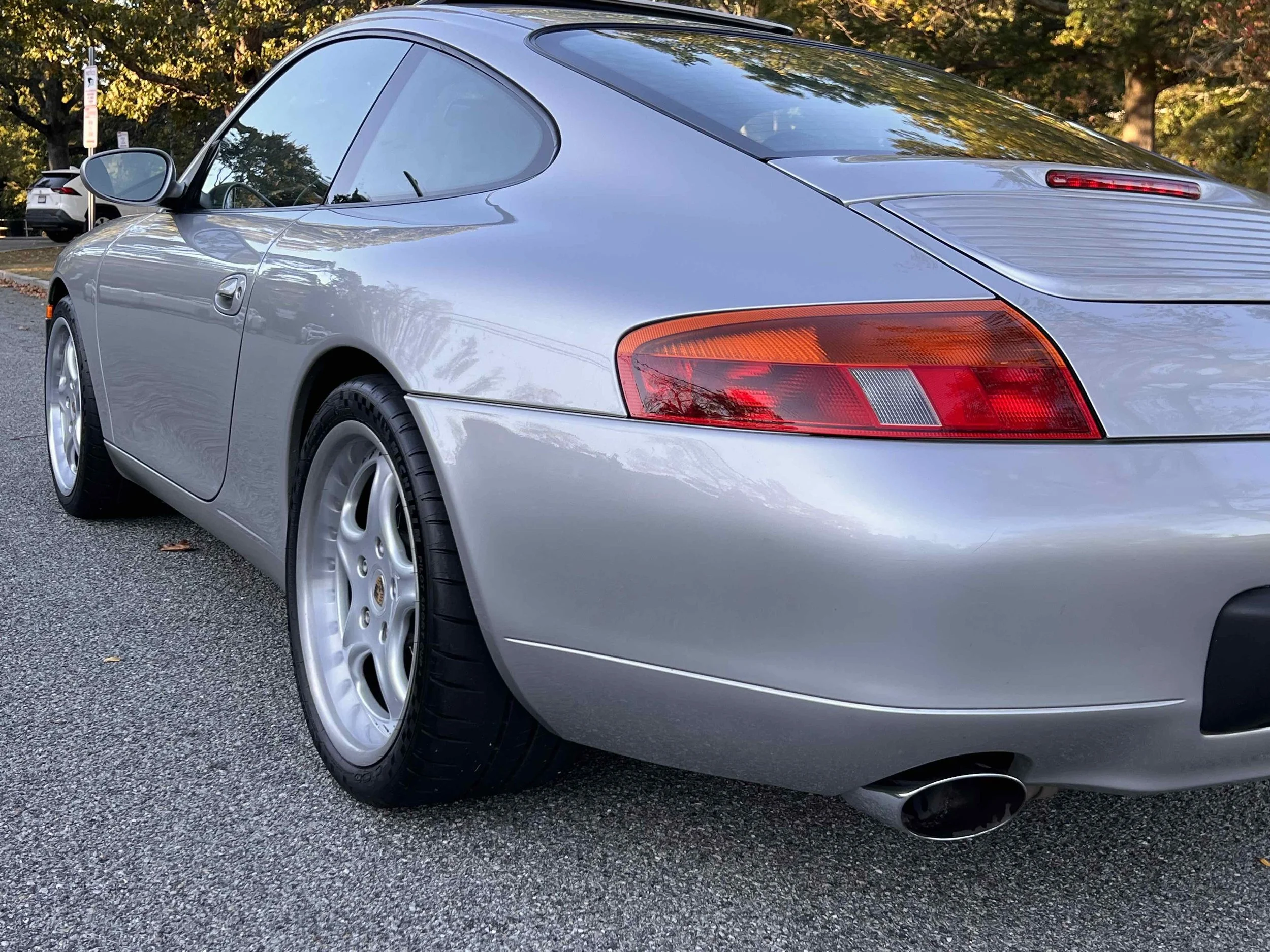 Rear and passenger side of a silver Porsche 911 parked on an asphalt surface with trees in the background.