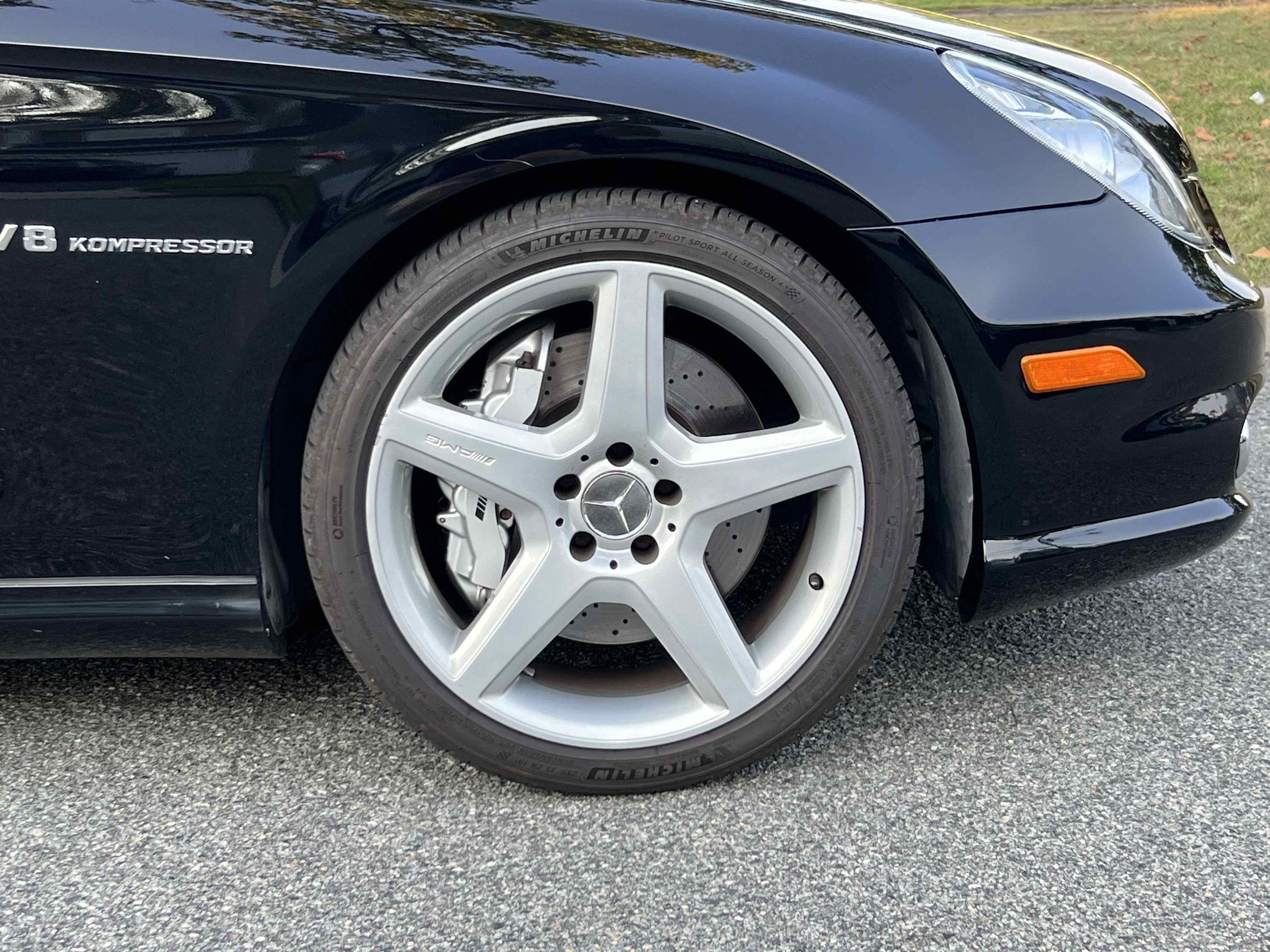 Close-up of the front wheel of a black Mercedes-Benz car, showing a silver AMG rim, brake caliper, and Michelin tire, parked on a paved surface with a grassy area in the background.
