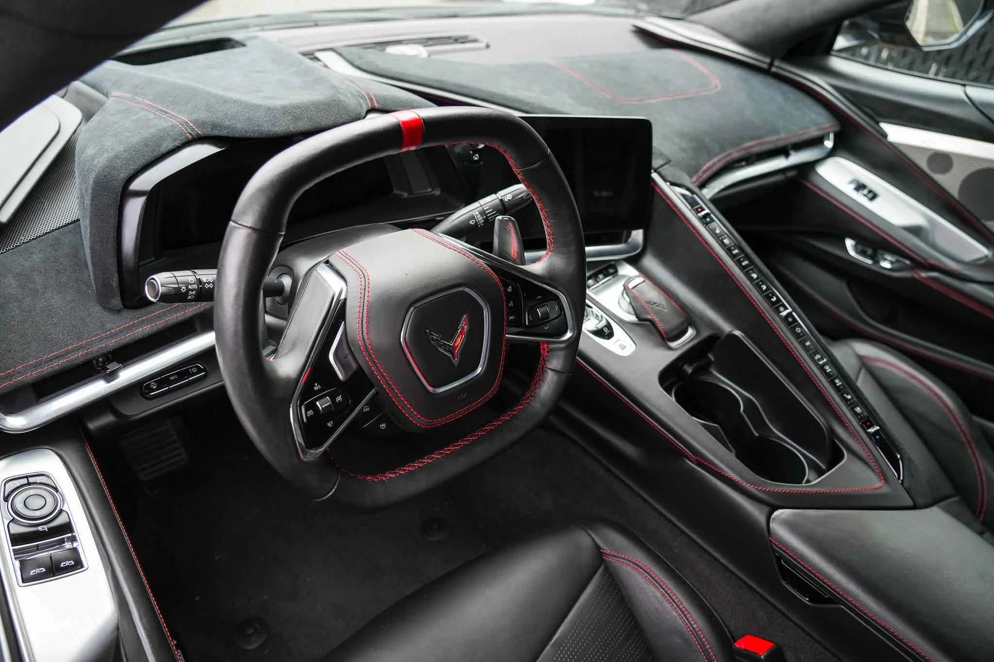 Interior of a modern sports car dashboard featuring a leather-wrapped steering wheel with red stitching, a digital display, cup holders, and control buttons.