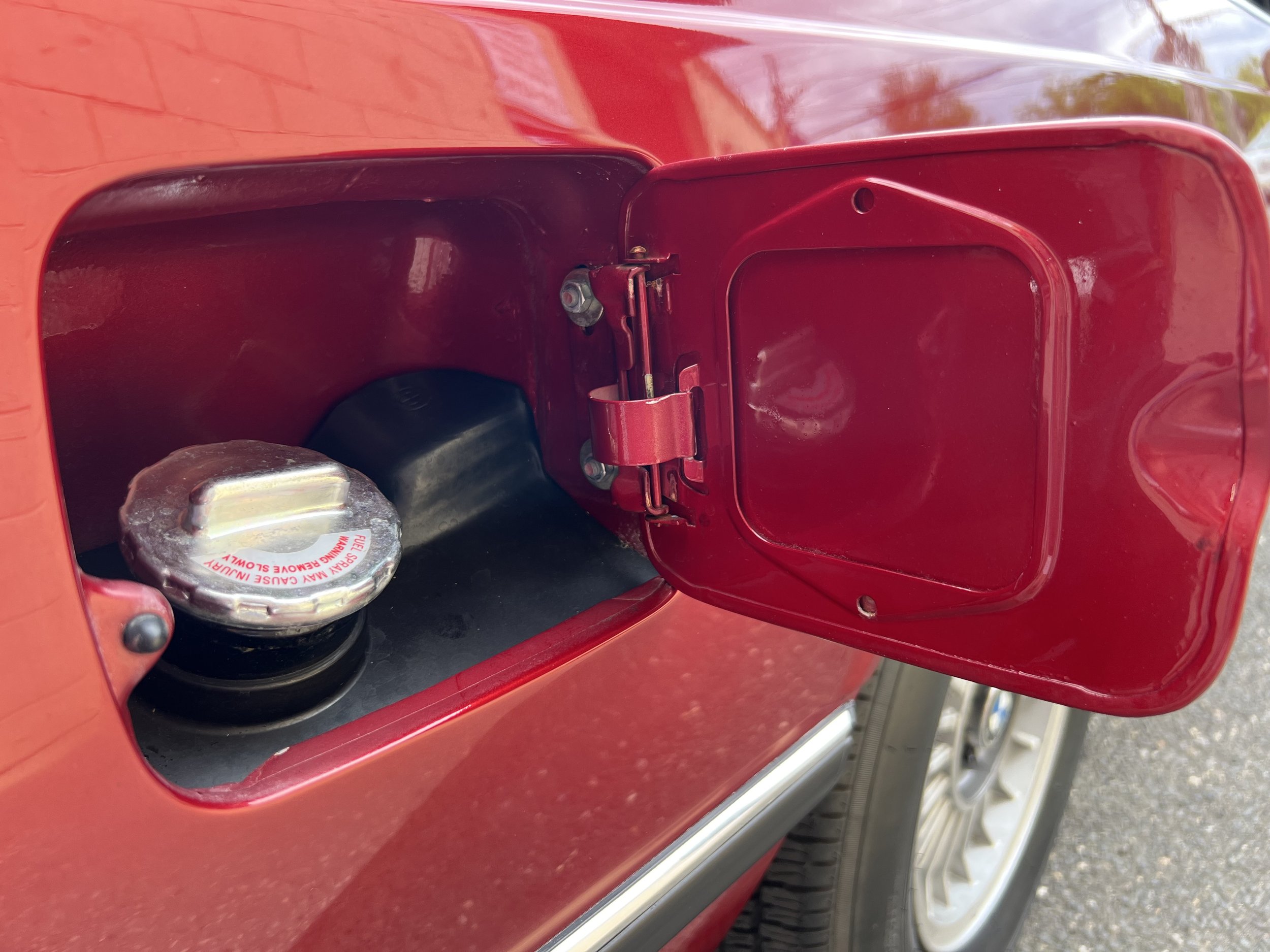 Open fuel tank door showing fuel cap and black fuel inlet on a red 1977 BMW 630 csi car.