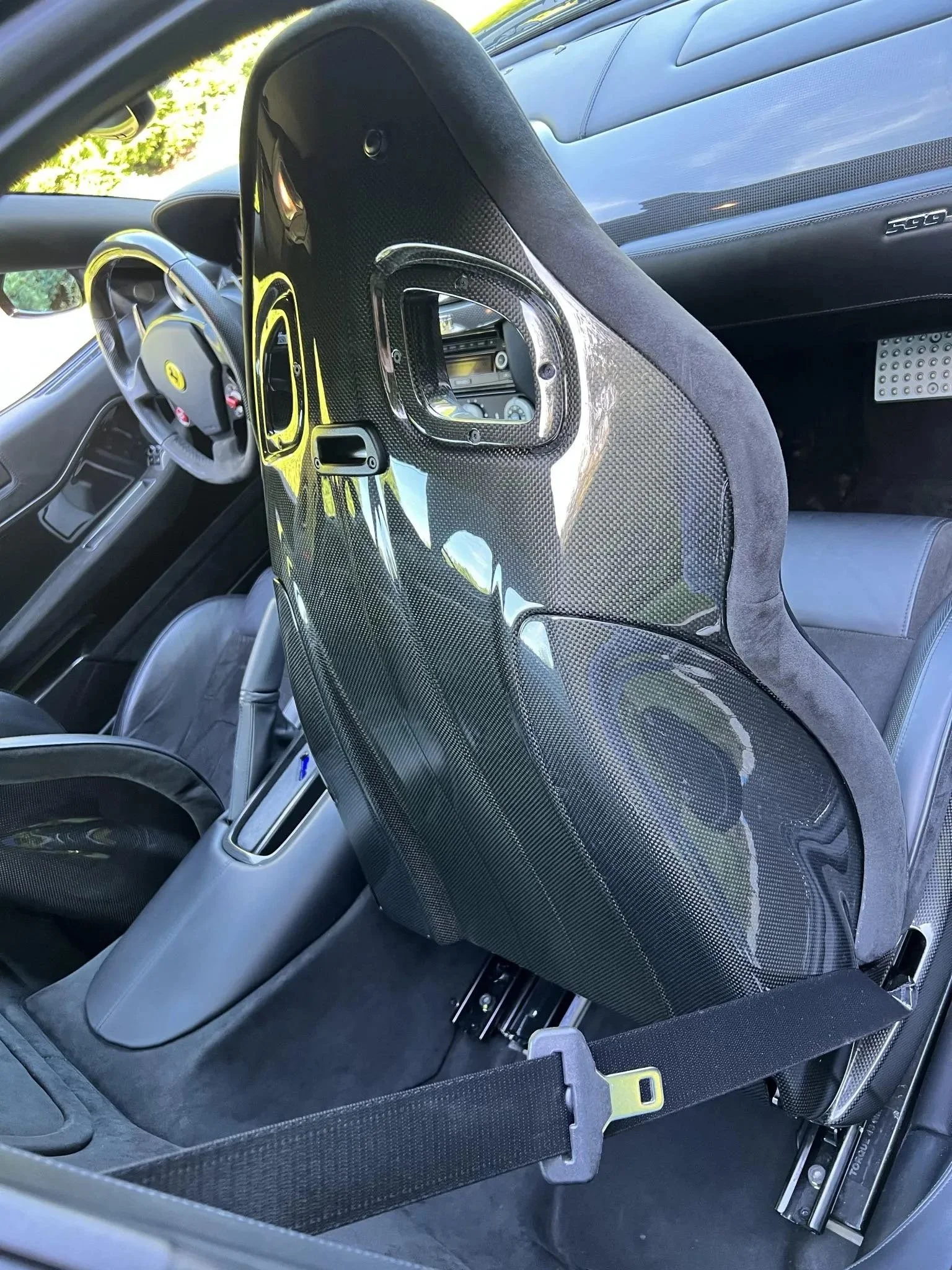 Interior view of a racing car showing a carbon fiber racing seat with a harness, steering wheel, and dashboard.