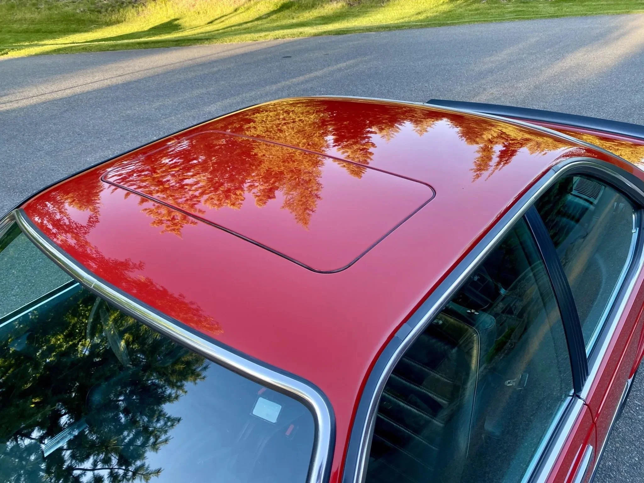 Red car with a sunroof, reflecting trees and blue sky, parked on a paved road with greenery in the background.