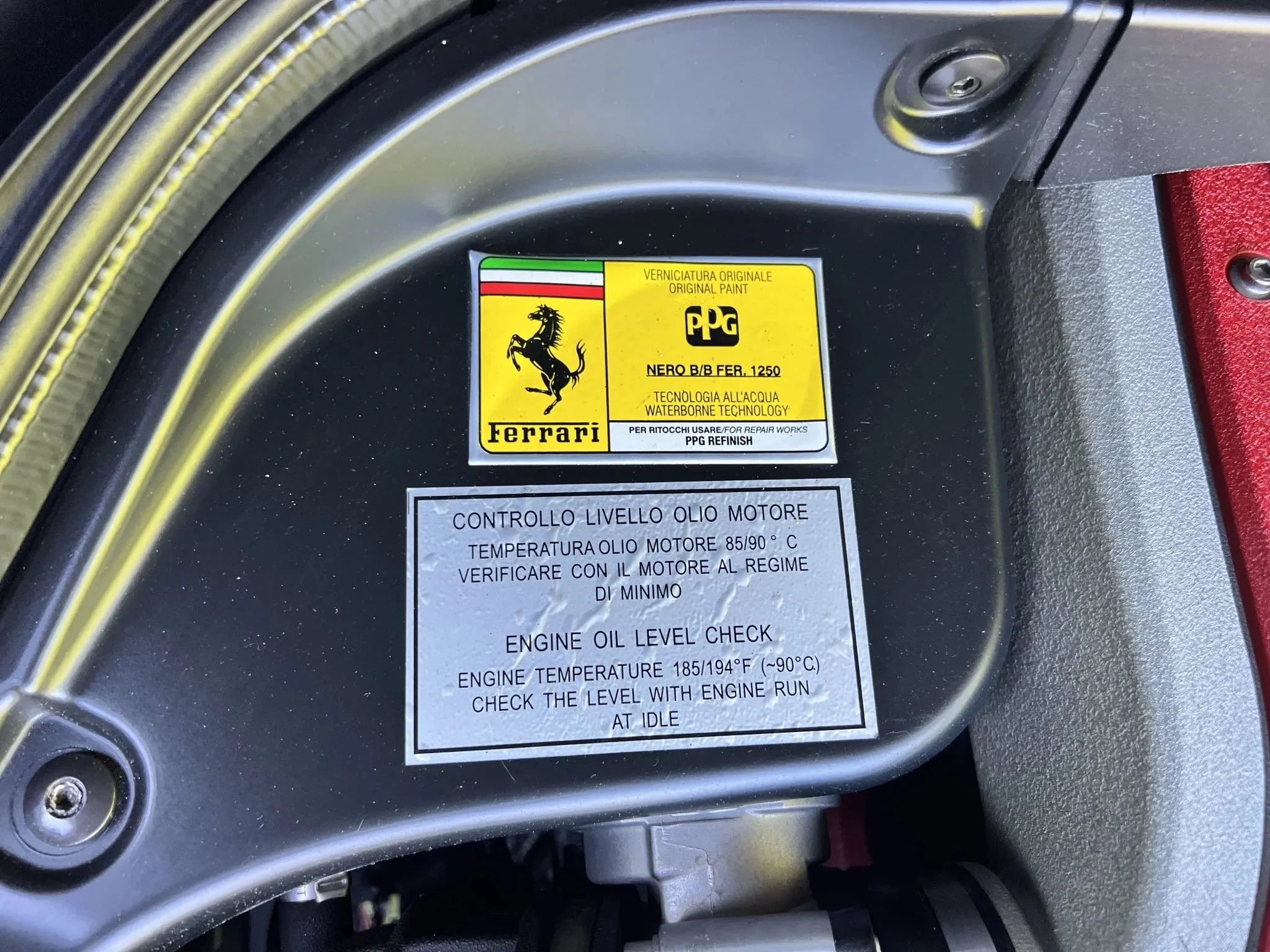 Close-up of a Ferrari engine compartment with labels indicating oil levels and original paint, including a Ferrari logo and a waterborne technology label.