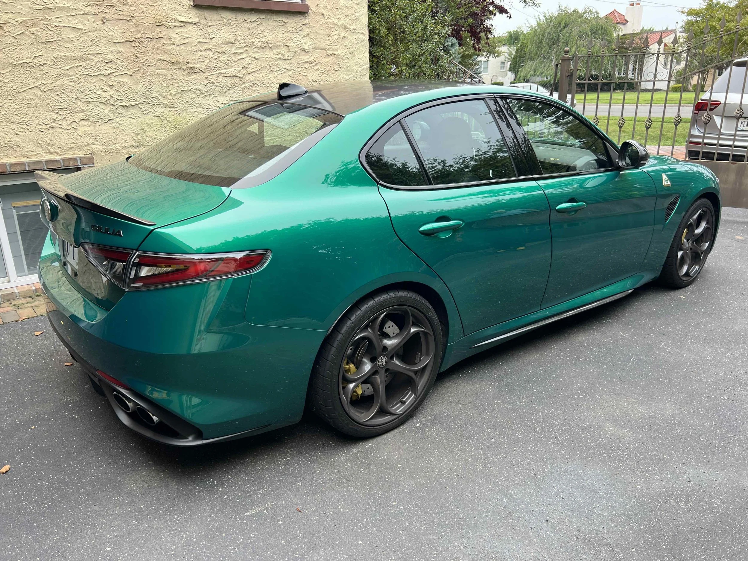 A teal-colored Alfa Romeo Giulia parked on a driveway next to a beige wall, with a black spoiler, dark alloy wheels, and quad exhaust pipes.