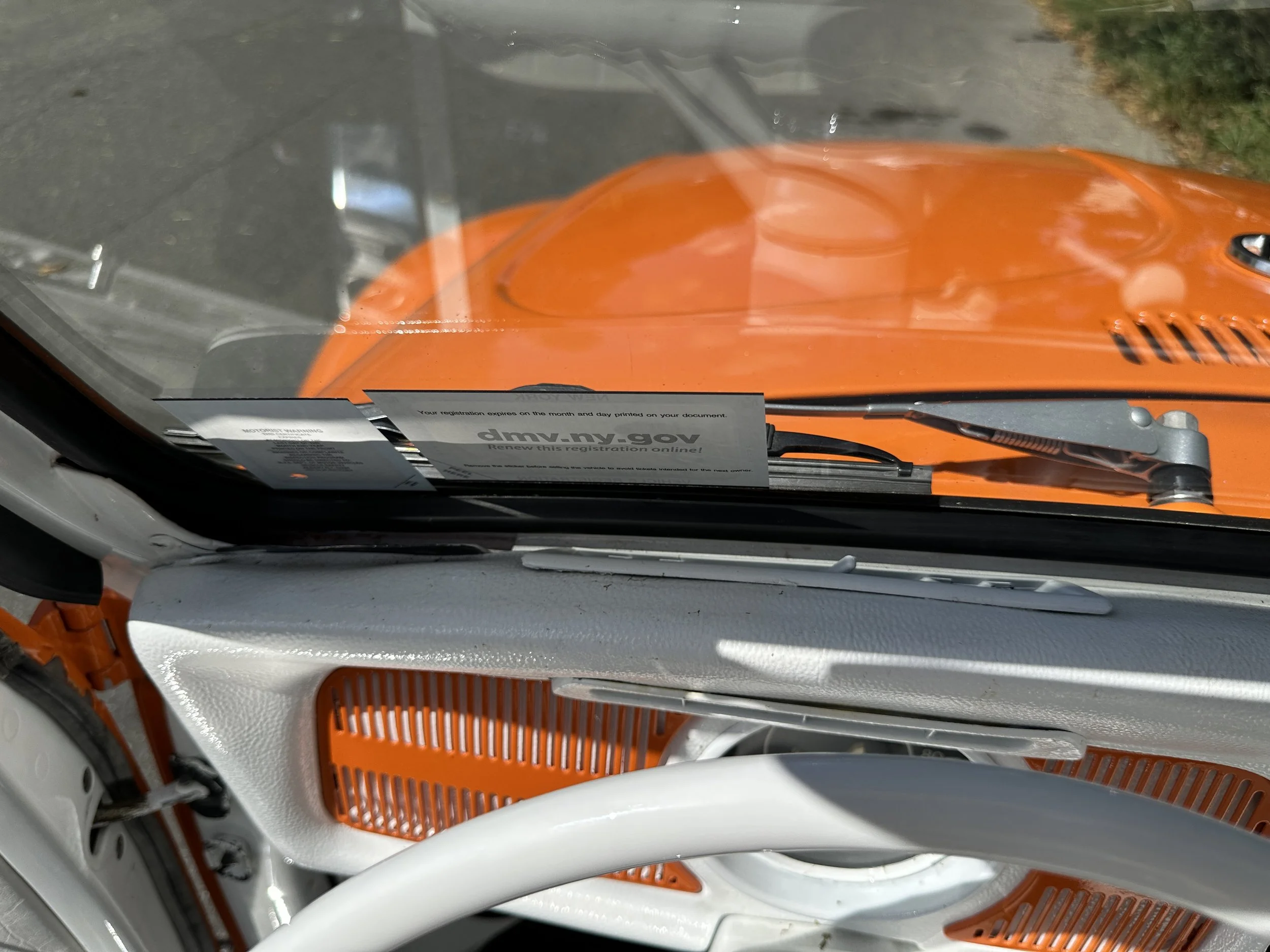 View from inside a vehicle showing the dashboard and windshield. The vehicle is orange with an orange hood visible through the windshield. There is a parking ticket on the dashboard.