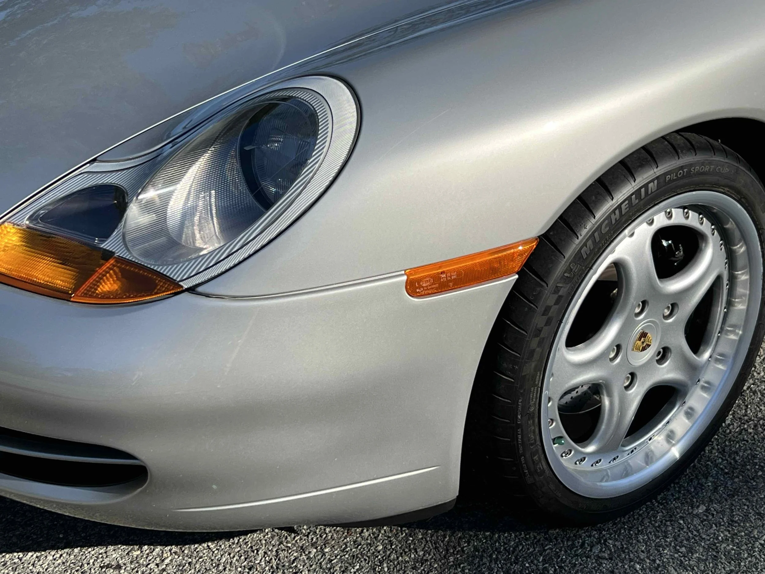 Close-up of the front left side of a silver Porsche sports car, showing the headlight, tire, and part of the bumper.
