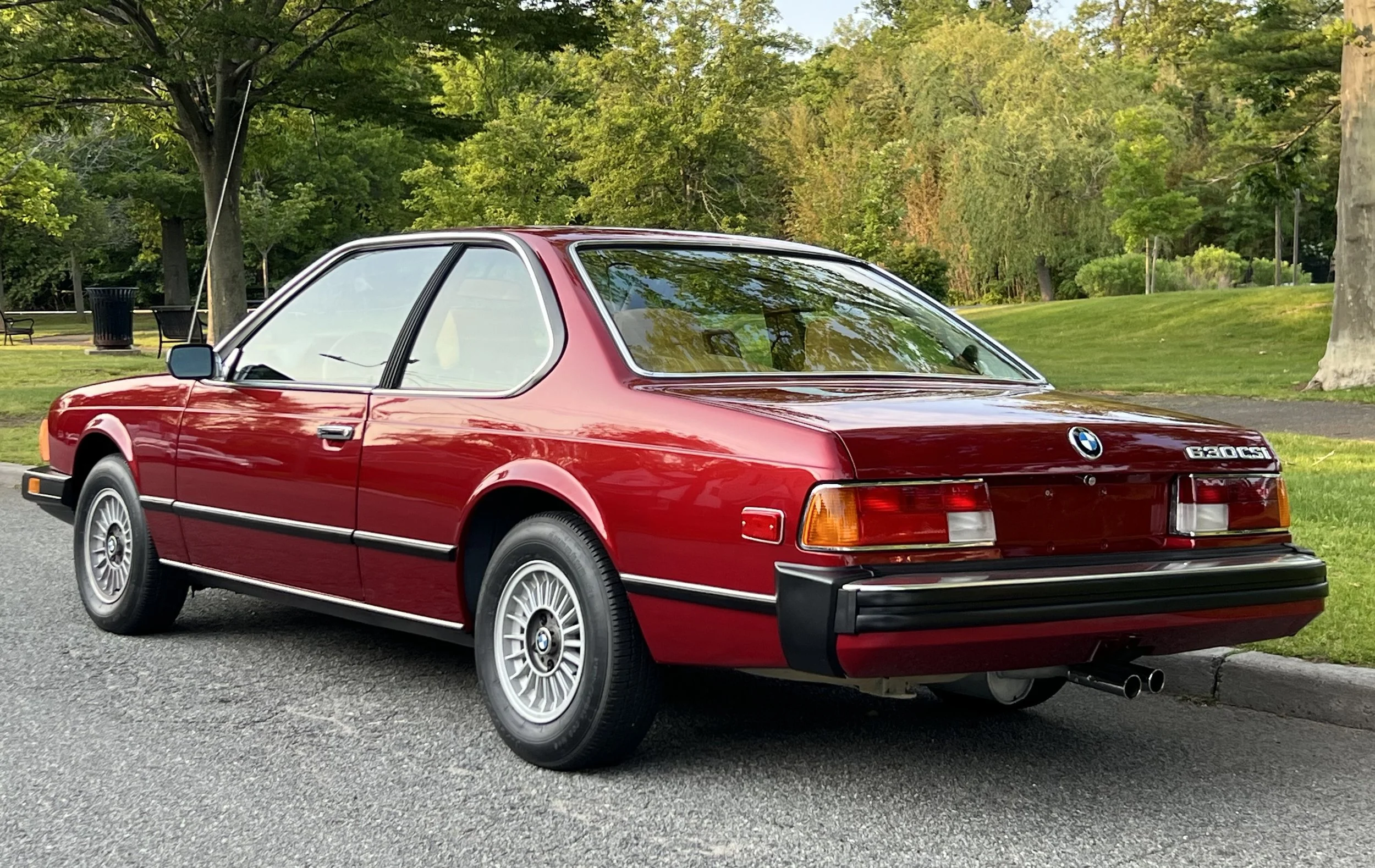 Ruby Red 1977 BMW 630 csi on display in park with trees, grass, and lake in background for sale at FlowHaus