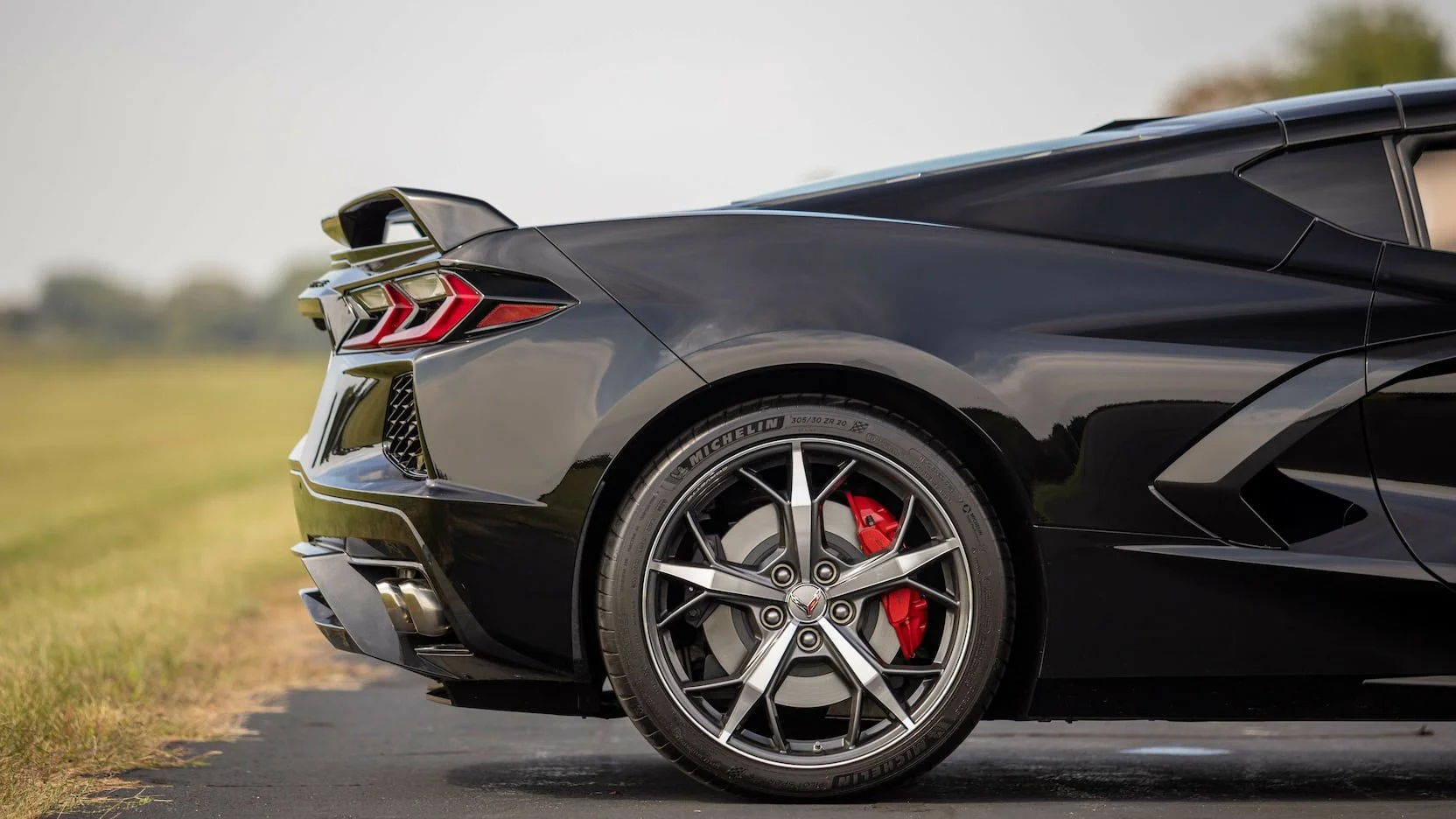 Close-up of the rear section of a sleek black sports car with red brake calipers and Michelin tires, parked on a paved surface with grass and trees in the background.