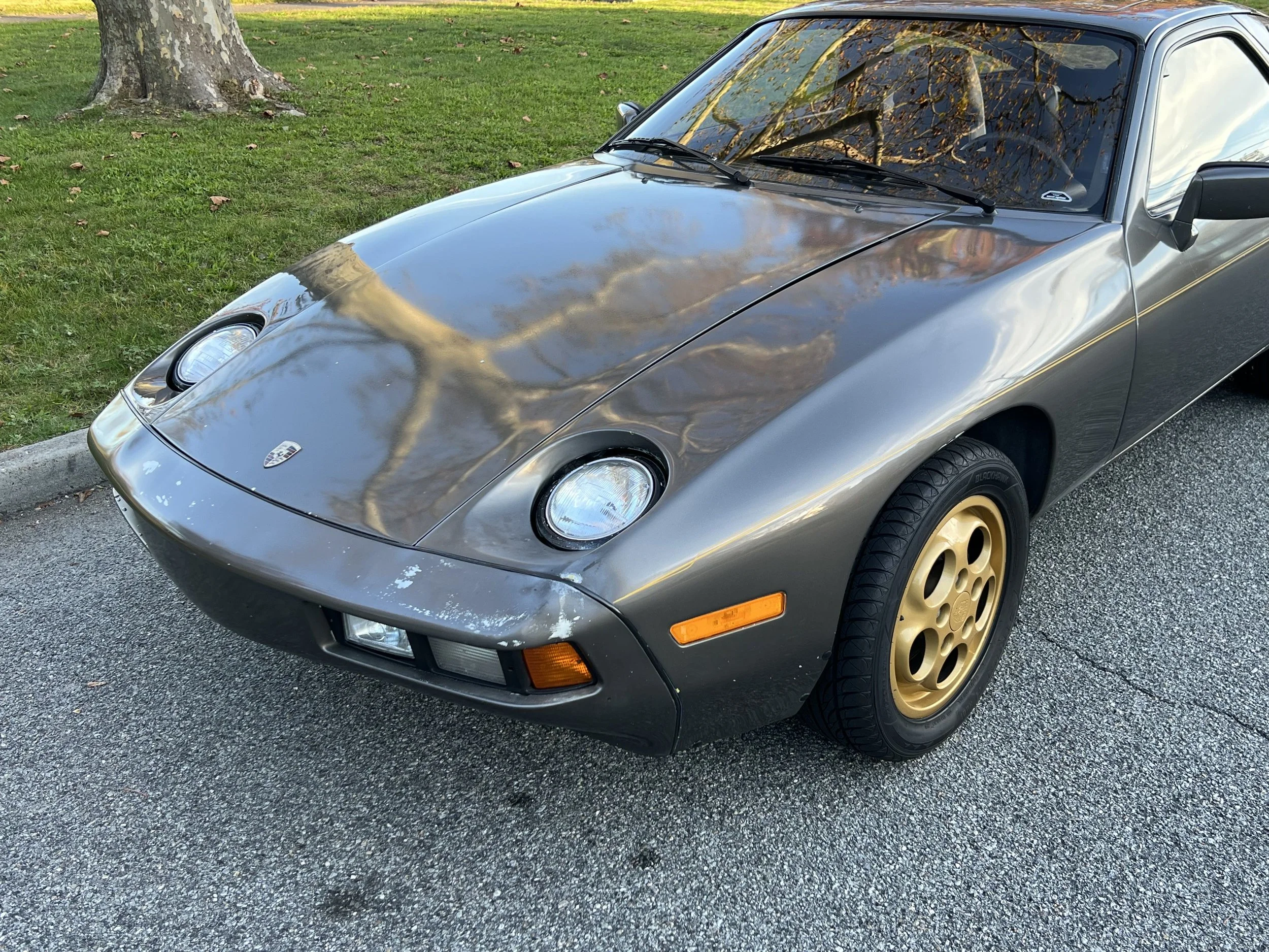 A vintage gray Porsche sports car with gold wheels parked on a street, with a tree and grass in the background.