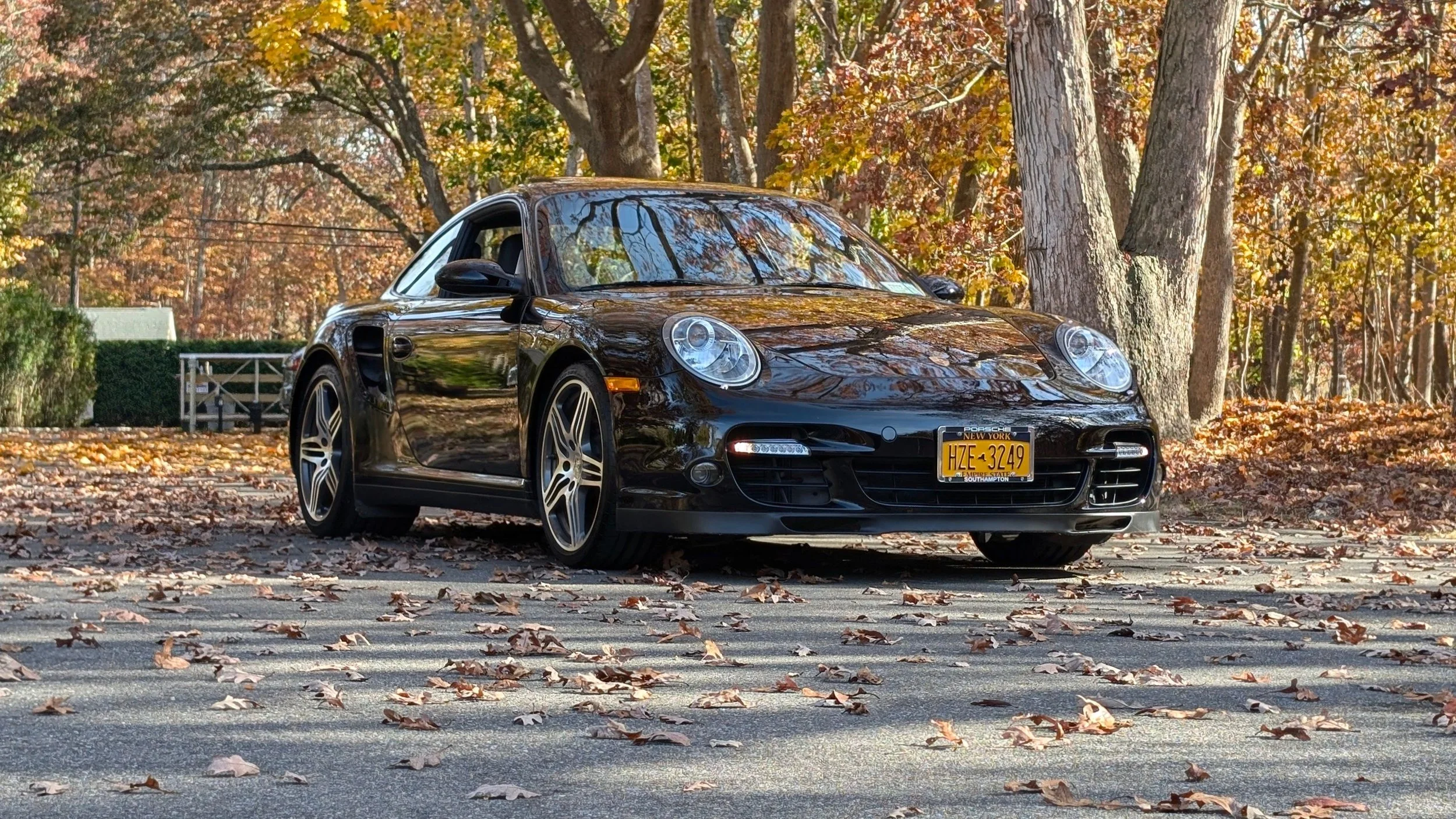 A black Porsche sports car parked on an autumn leaf-covered road amid trees with fall foliage.