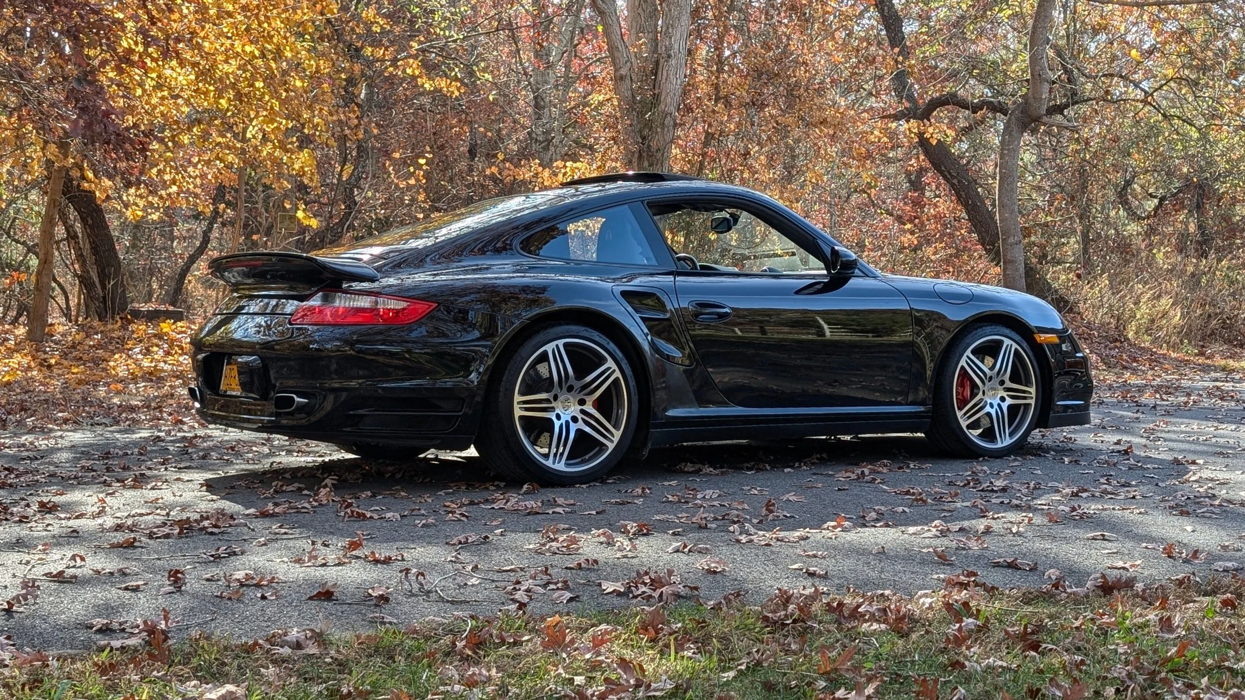Black sports car parked on a leaf-covered road with autumn trees in the background.