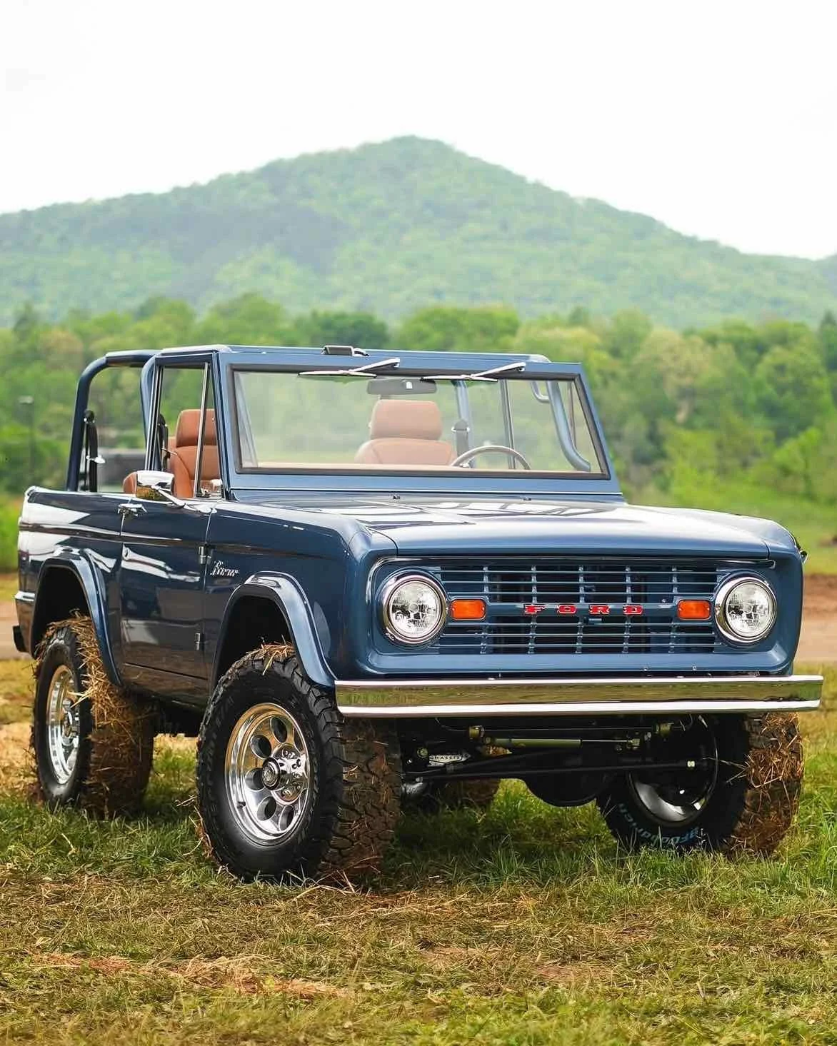 A vintage blue Ford Bronco off-road vehicle with tan interior and large tires, parked on grassy terrain with a mountain and green trees in the background.