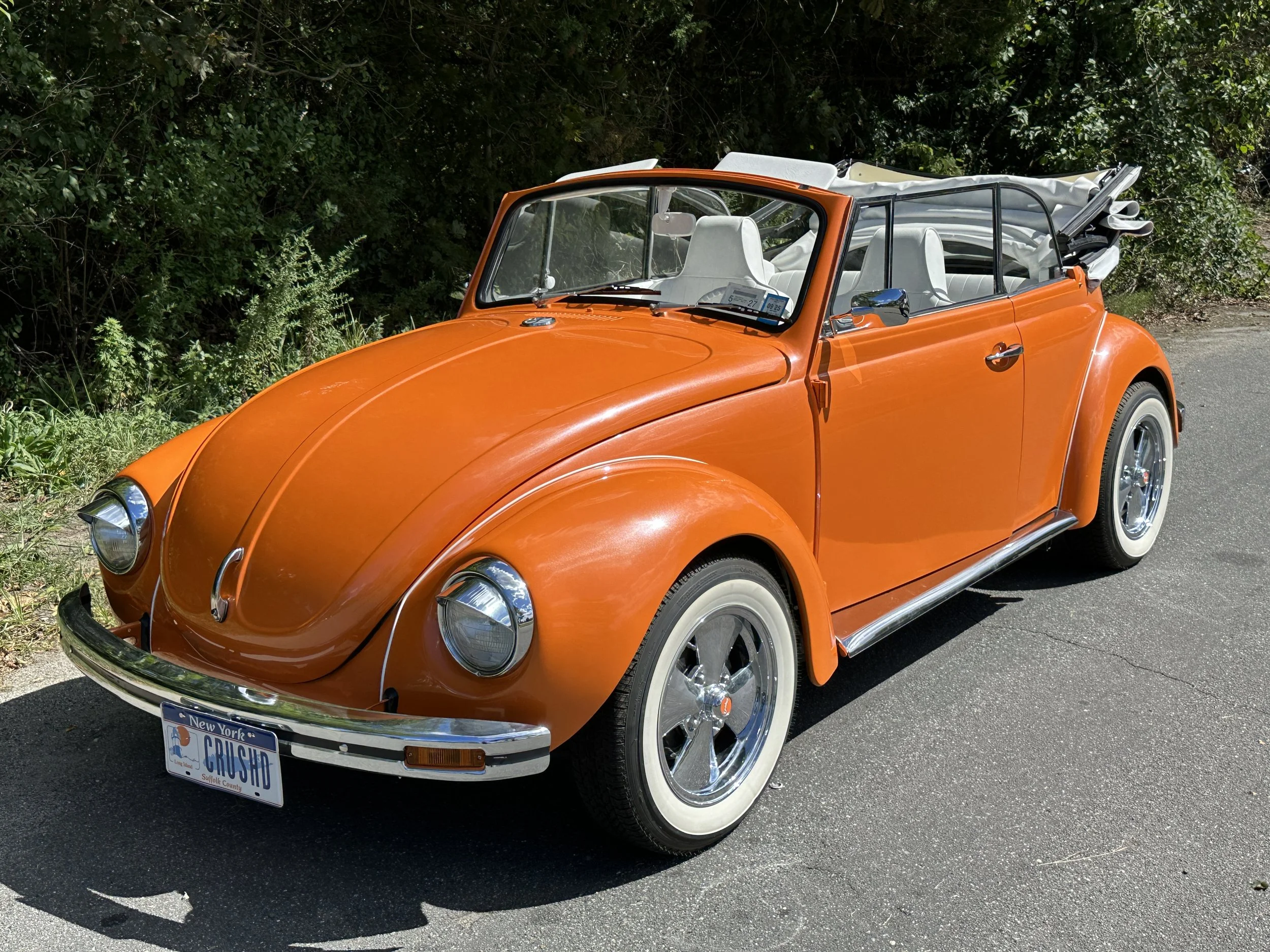 An orange vintage convertible Volkswagen Beetle with white interior and a white folded soft top, parked on a paved road beside green bushes.