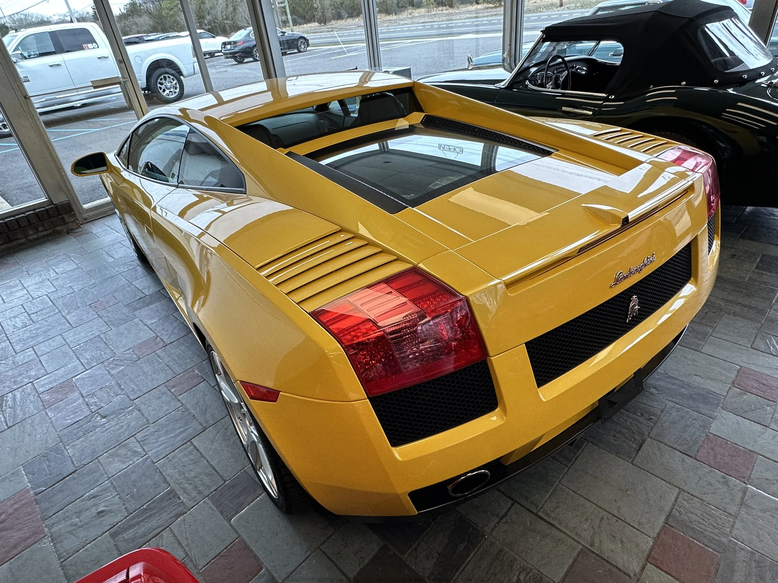 Yellow Lamborghini Gallardo sports car on display inside with a black convertible car next to it. Large windows in background showing a parking lot outside.