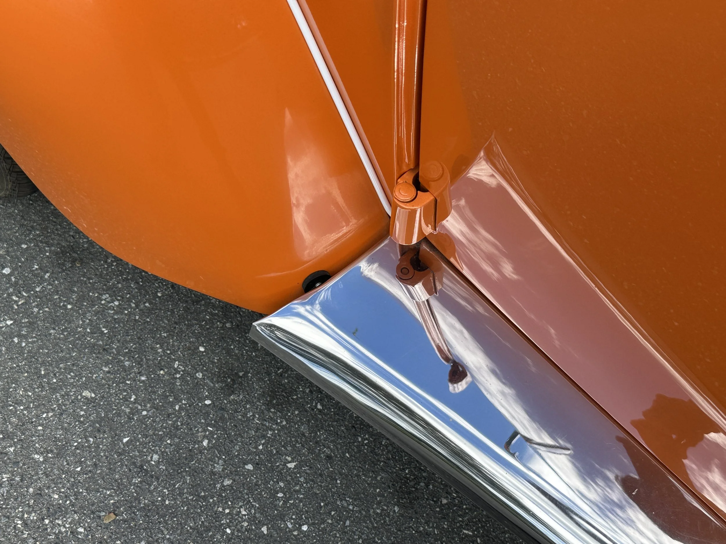 Close-up of the corner of an orange vintage vehicle, showing part of the wheel well, chrome bumper, and door hinge.