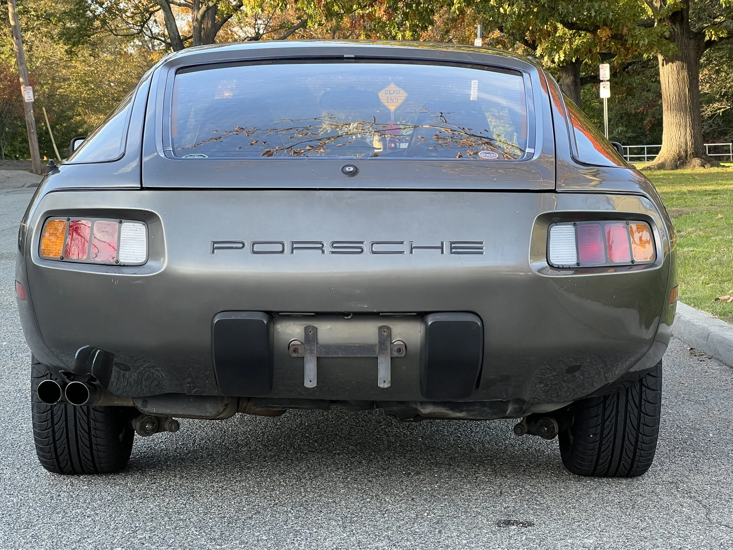 Rear view of a gray classic Porsche sports car parked on the street with trees and a small park in the background.