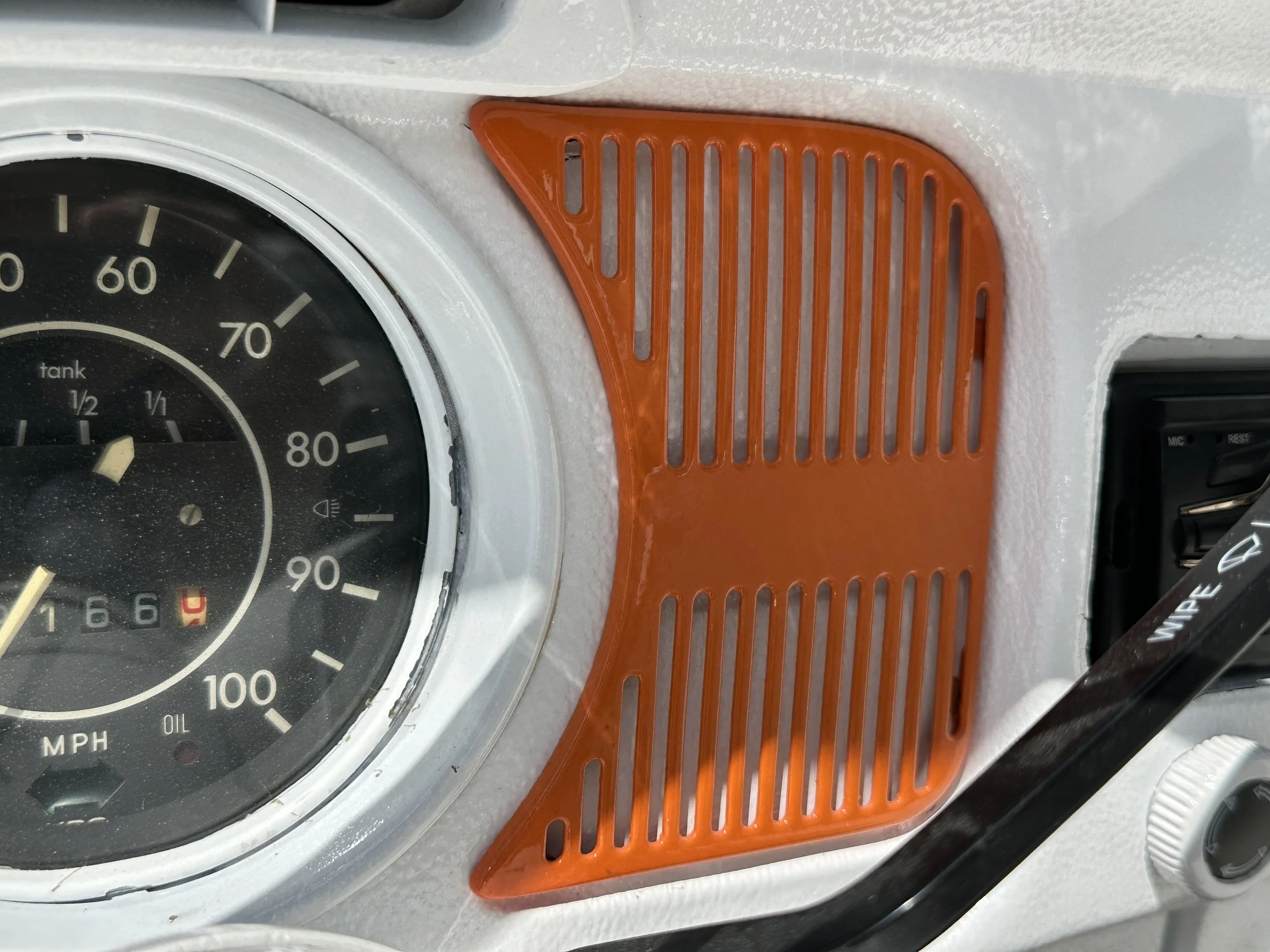 Close-up of a vehicle dashboard showing a speedometer, orange vent cover, and part of an air vent with control knobs.
