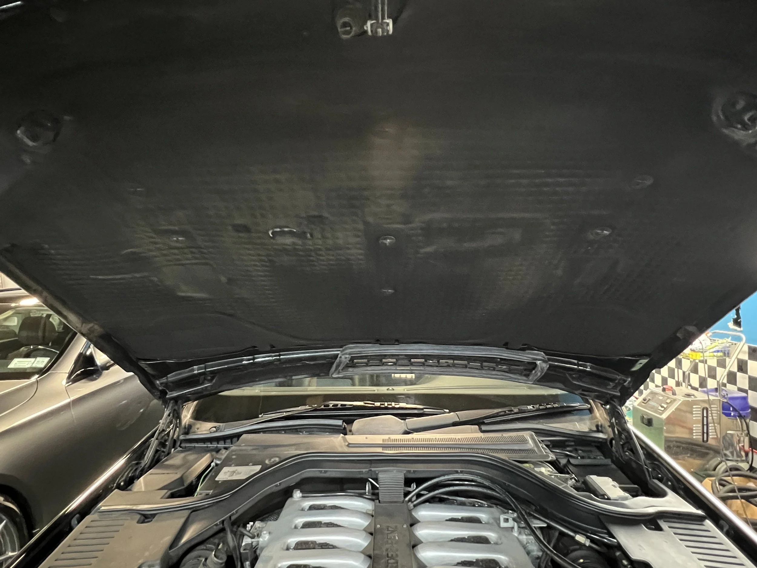 View under open car hood showing engine bay and components in a garage setting.