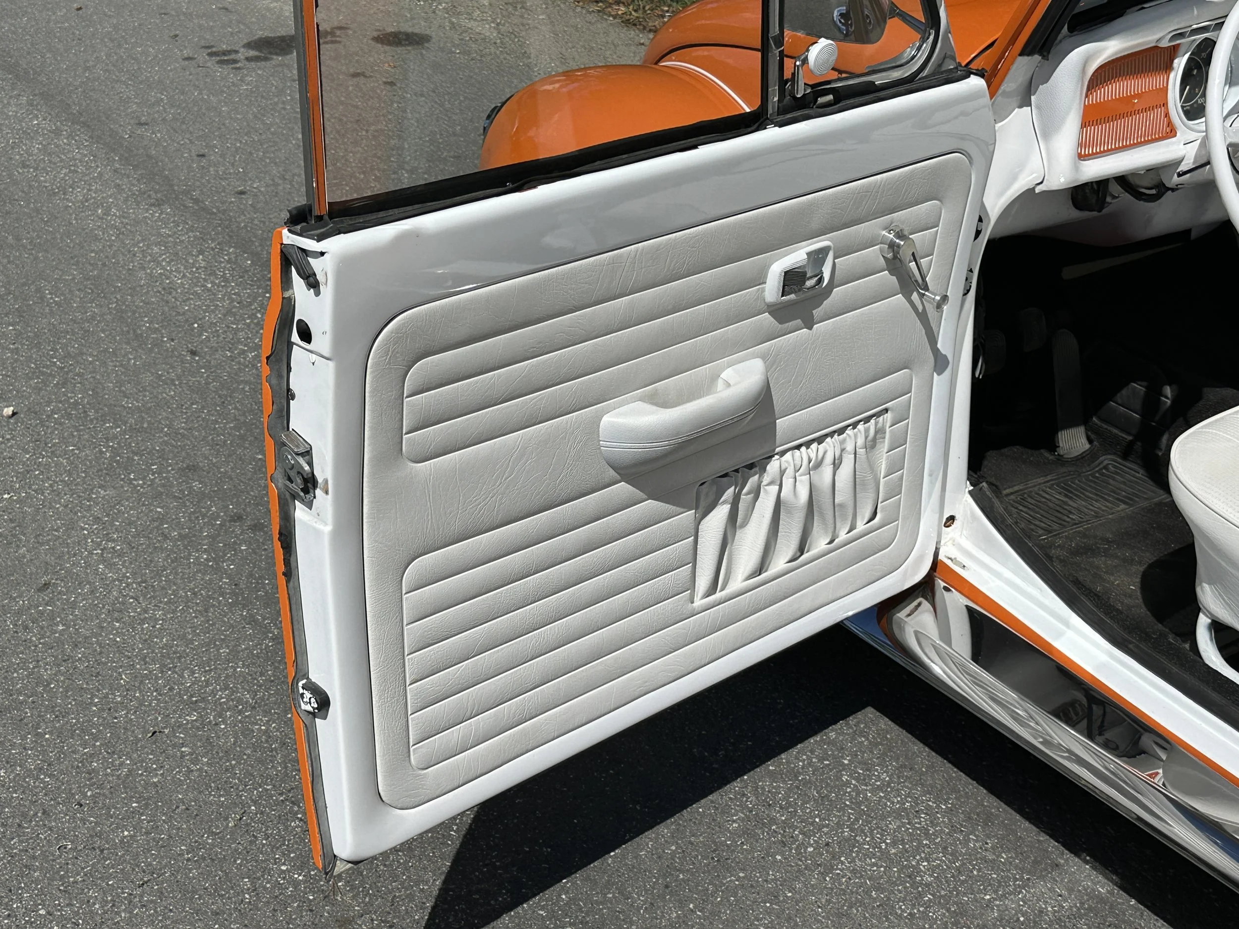 Close-up of a white vintage car door with a handle, window crank, and small storage pocket with elastic. The car's interior is black, and the exterior features orange accents.