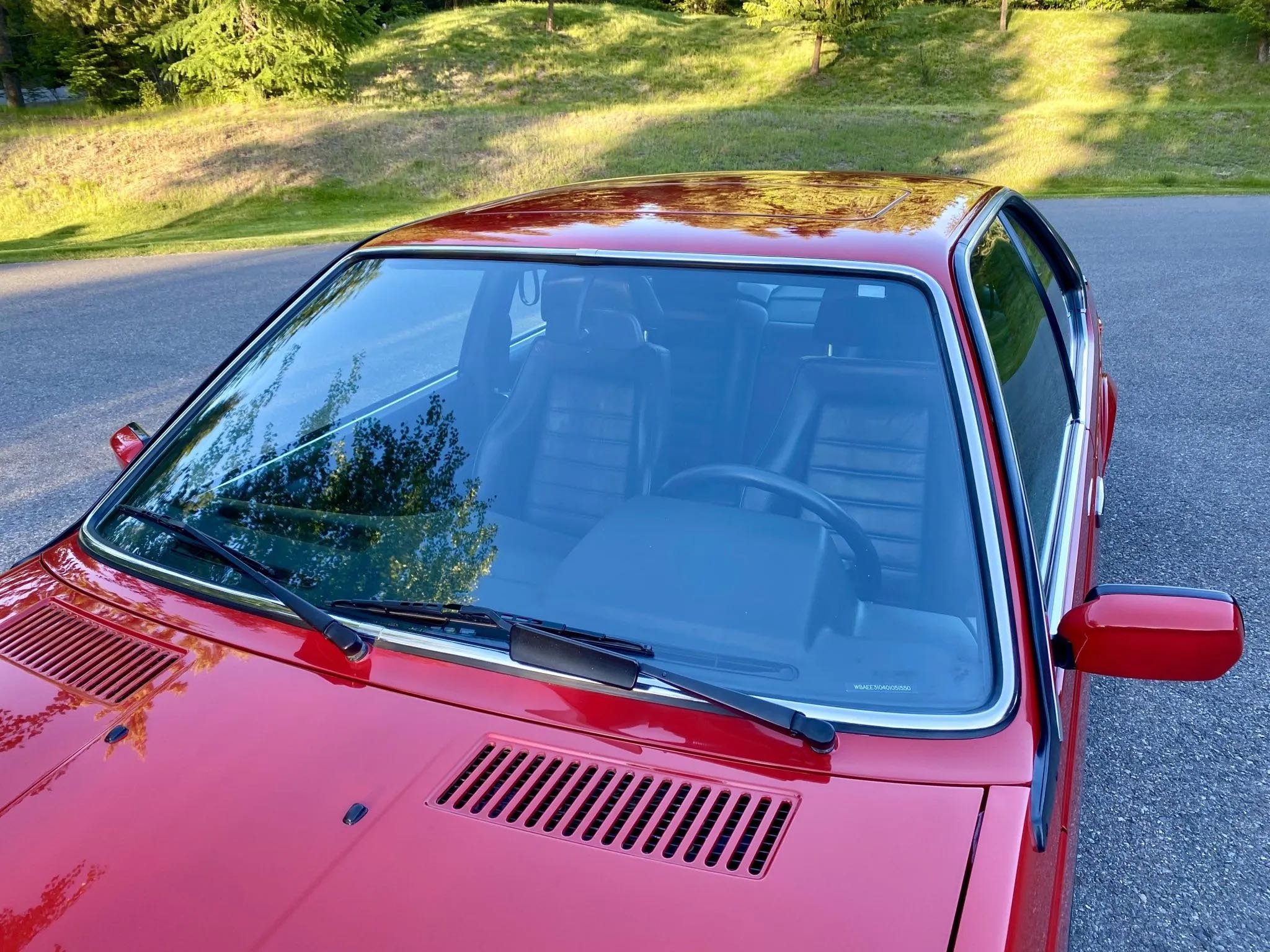 Red vintage car parked on a paved road with green grass and trees in the background, viewed from the front-top angle.