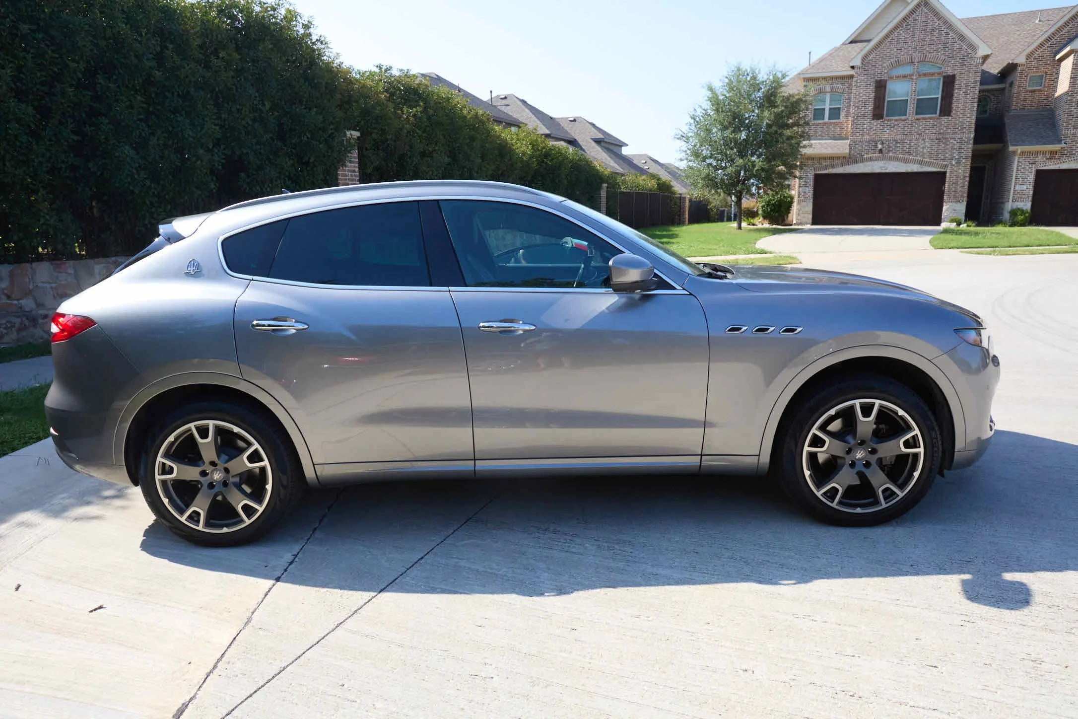 A silver luxury SUV parked on a driveway in front of a suburban house with brick exterior and a manicured lawn.