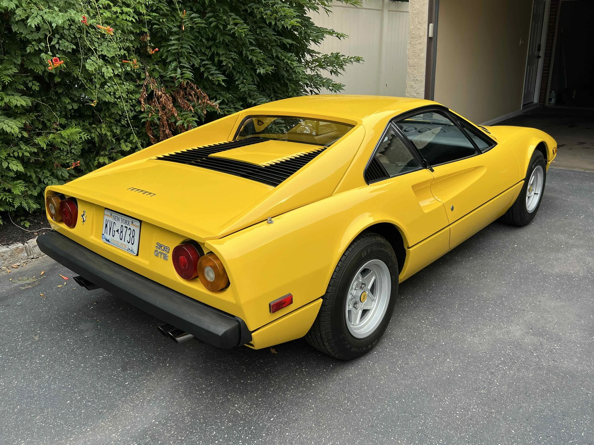 Yellow vintage Ferrari 308 GTB sports car parked on asphalt driveway near garage and green bushes.
