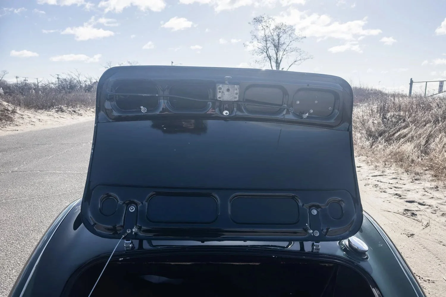 Close-up of the front trunk of a black vintage car with the hood open, showing the internal panel and hardware, outdoors on a sunny day with a clear sky and leafless trees in the background.