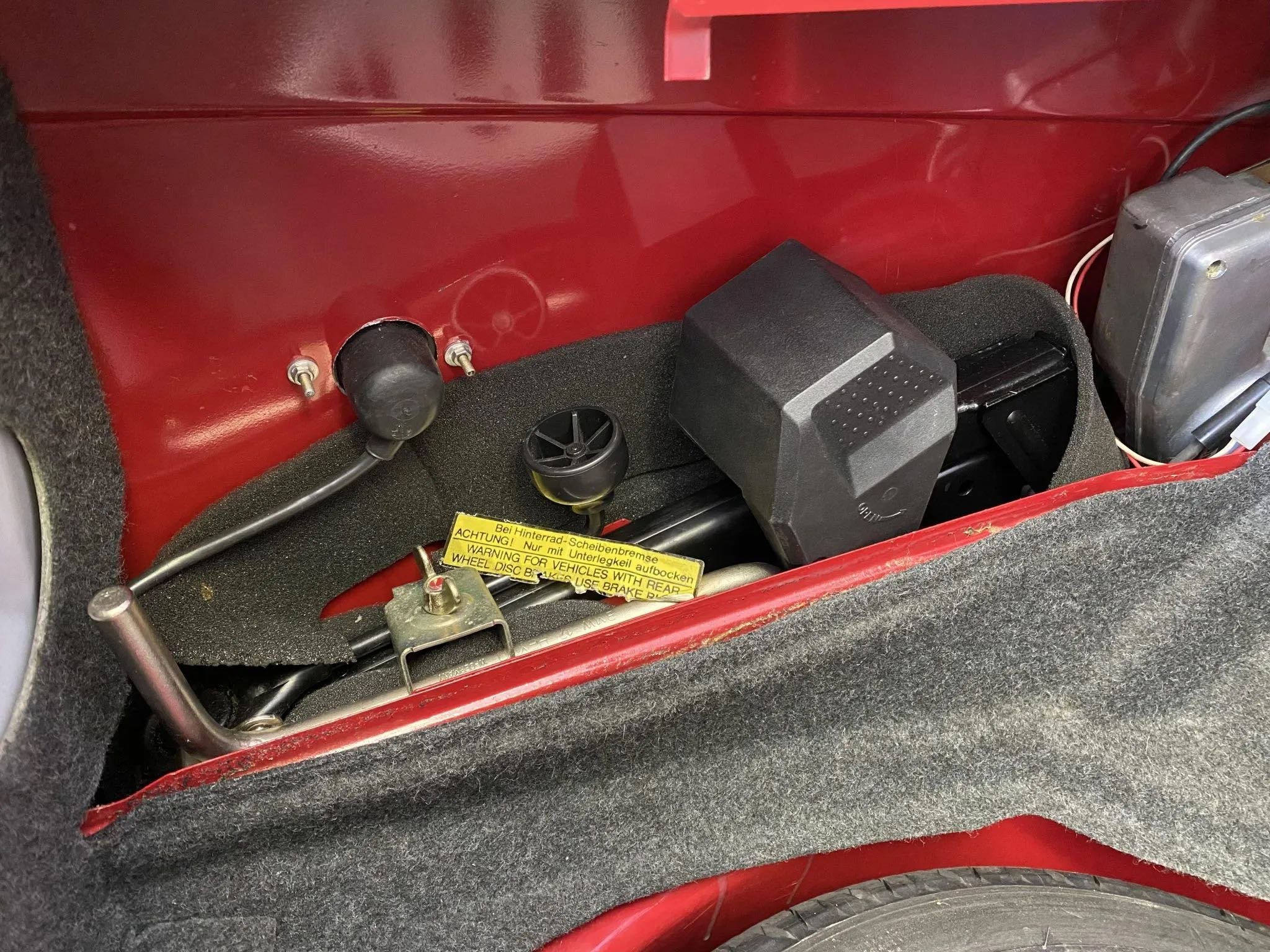 Interior of a classic red vehicle showing the rear wheel well area with various mechanical parts, wiring, and a warning label.