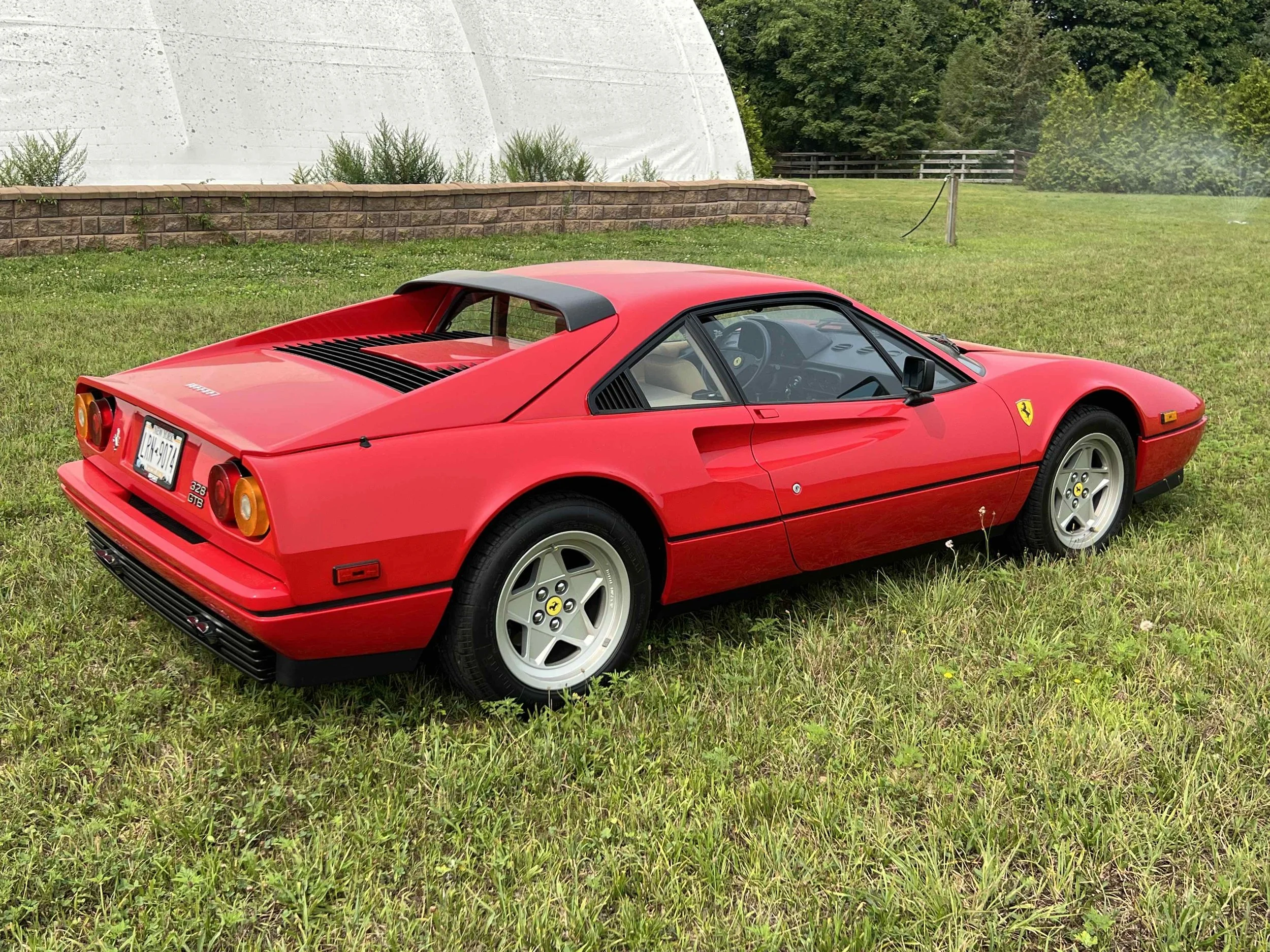 Red Ferrari 328 GTS parked on grass with green trees and a stone wall in the background.