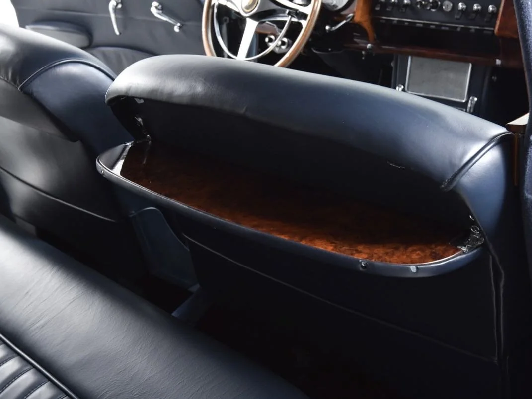 The interior of a vintage car showing a black leather seat and a wooden glove compartment with a cover open, revealing its inside.
