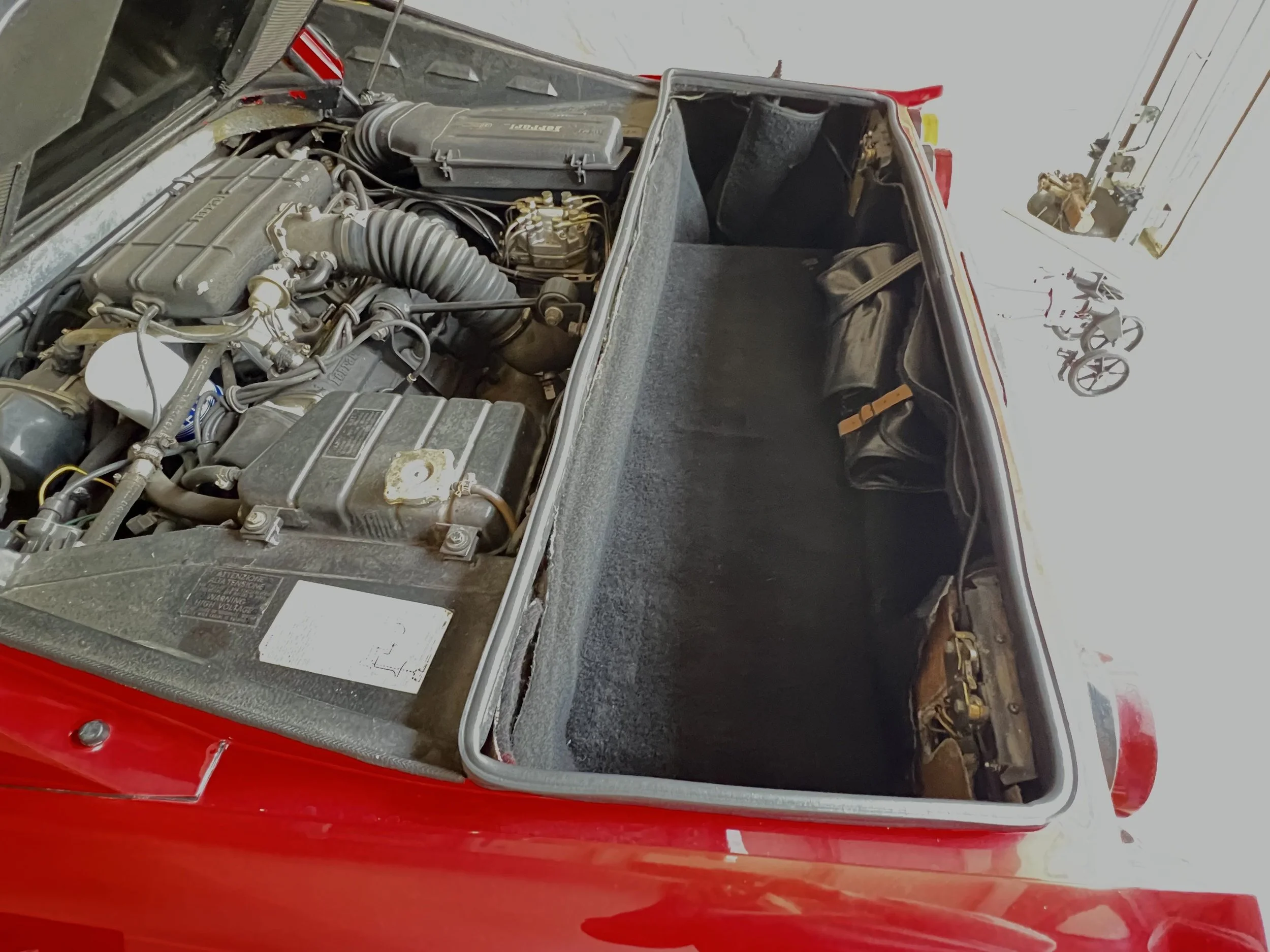 The image shows the open rear trunk of a red vintage sports car, revealing its engine compartment on the left and empty trunk space on the right.