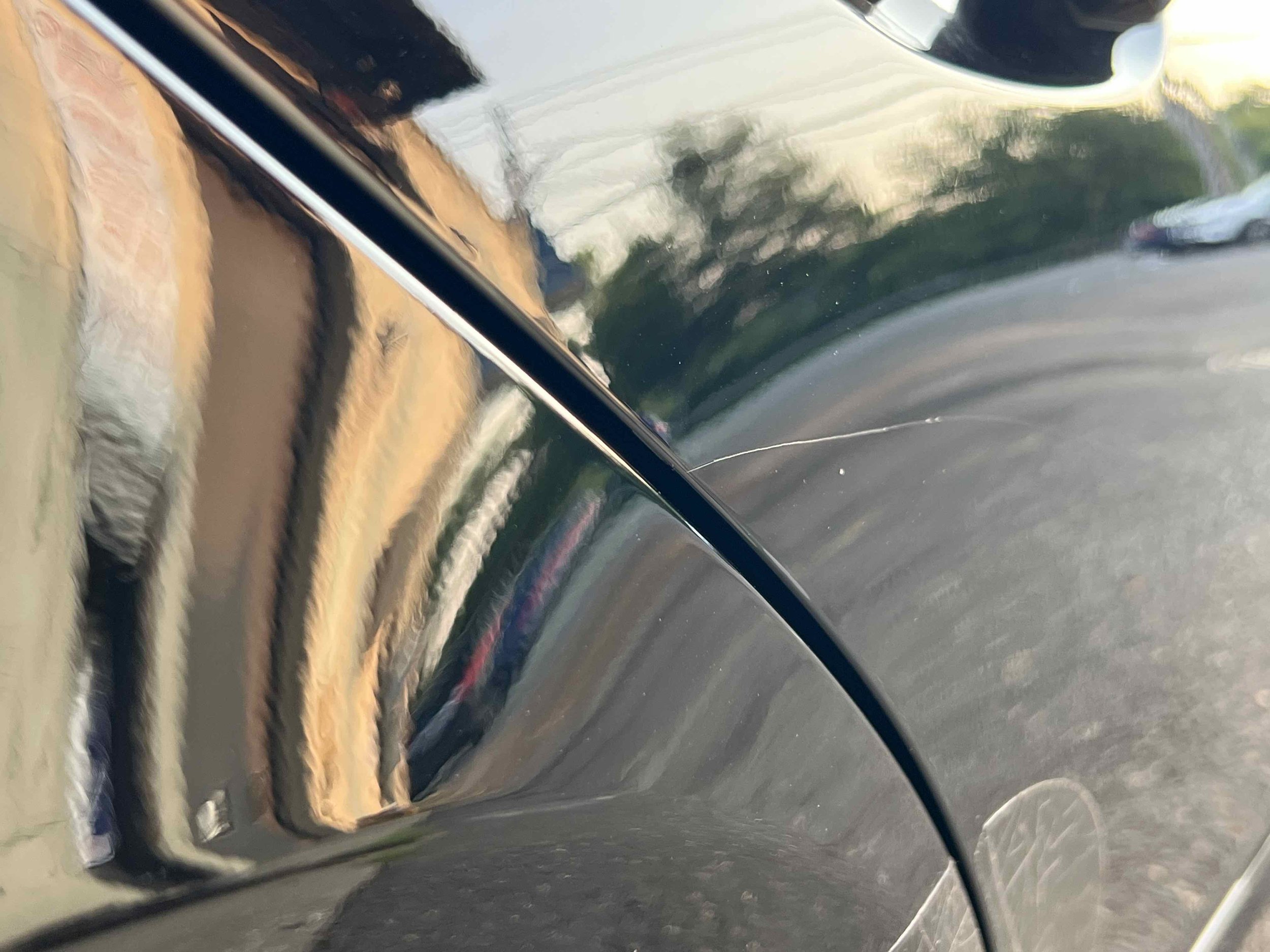 Close-up of a shiny black car reflecting trees, a building, and a white vehicle, with a visible scratch on the car's surface.