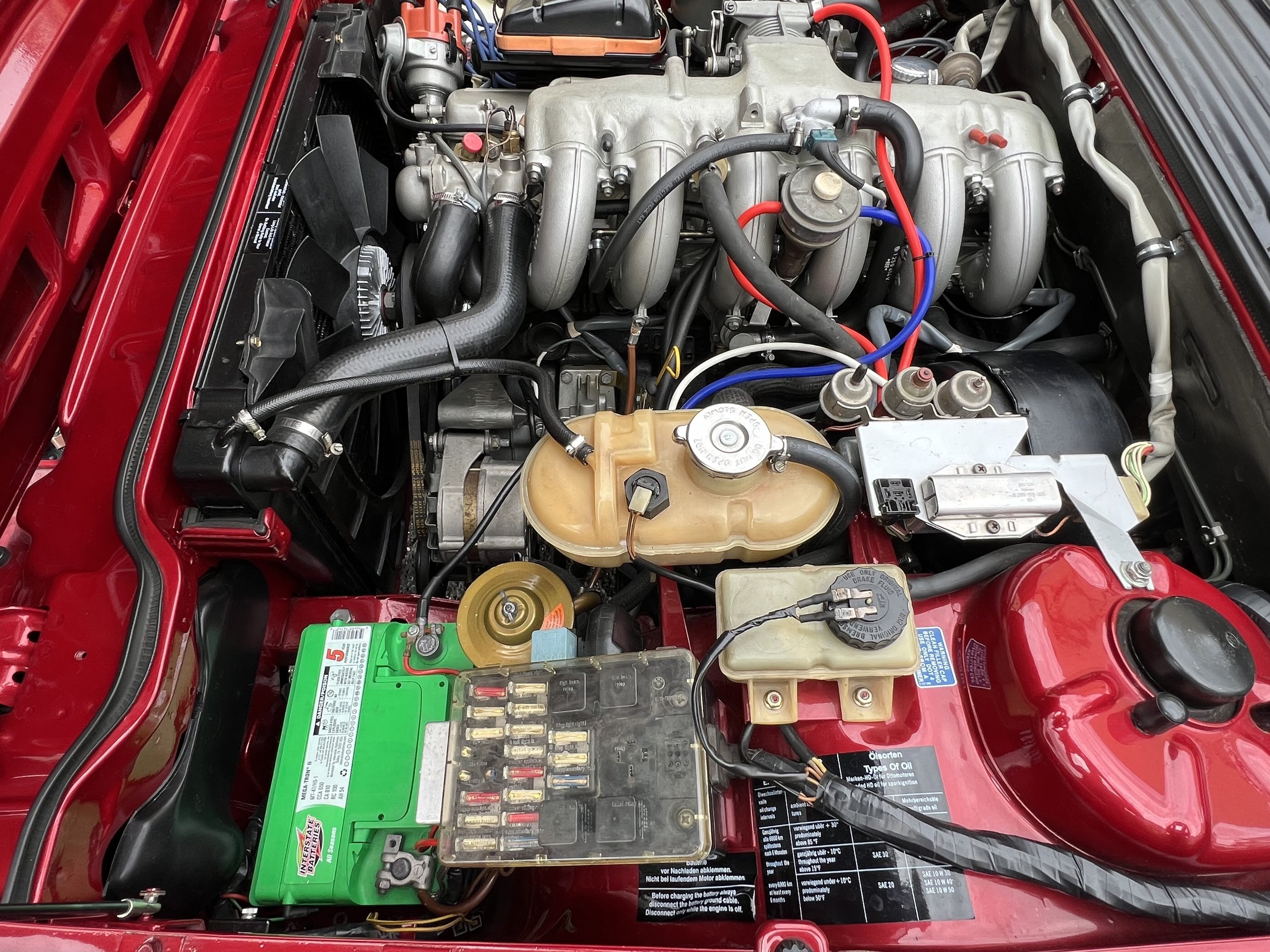 1977 BMW 630 csi car engine with various components, hoses, and a green battery in a red engine bay.