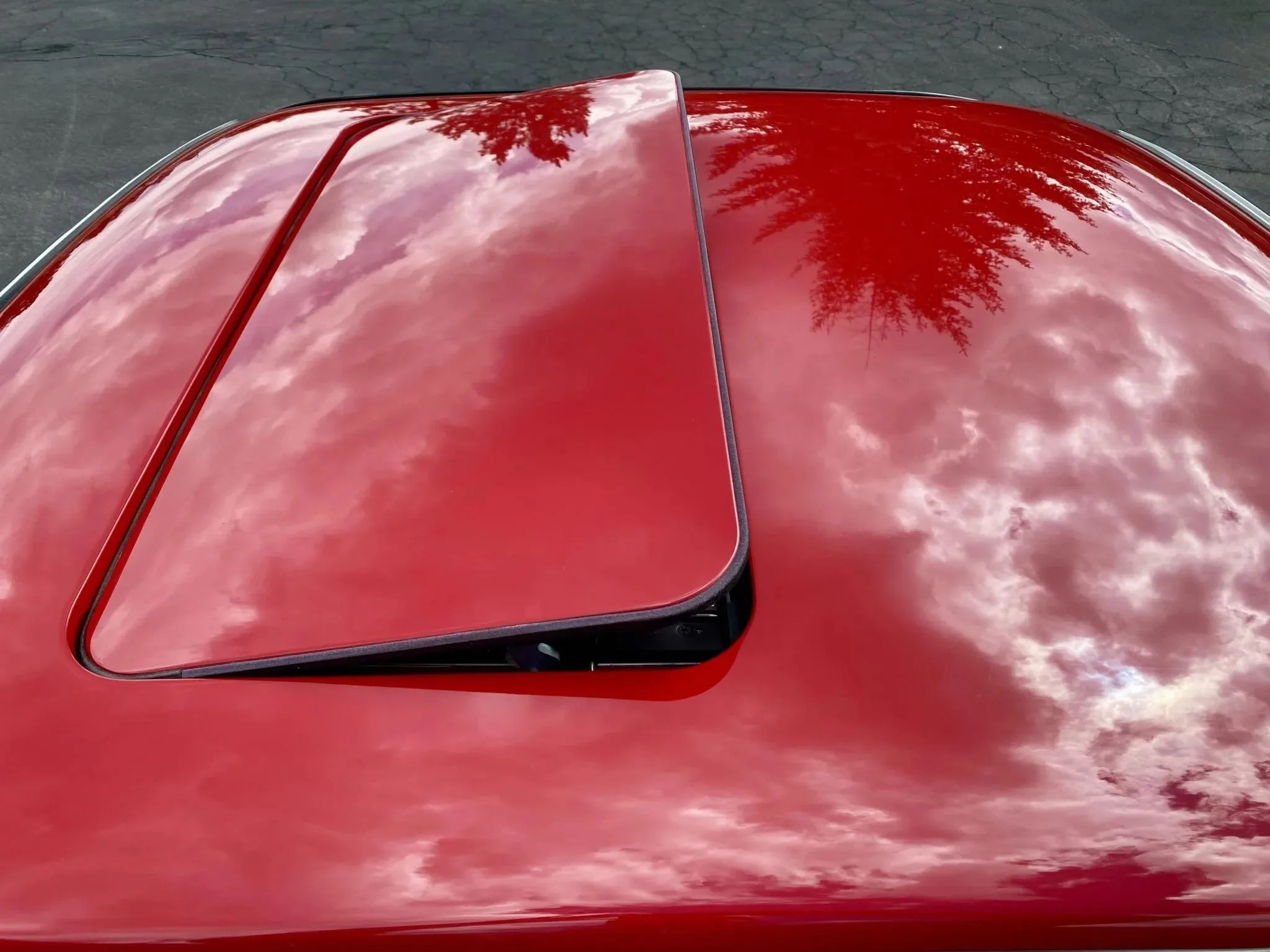 Close-up of a red car roof with a sunroof reflecting the cloudy sky and a tree.