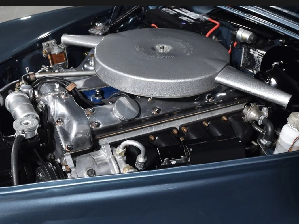Close-up of a vintage car engine with metallic components and an oval air cleaner cover on top.