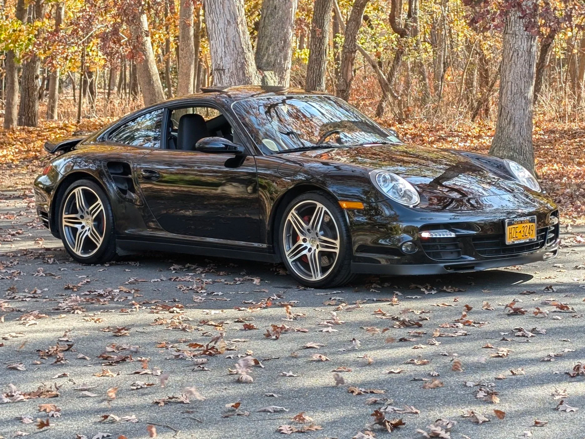 A black Porsche 911 convertible parked on a leaf-covered road in a forest with autumn foliage.