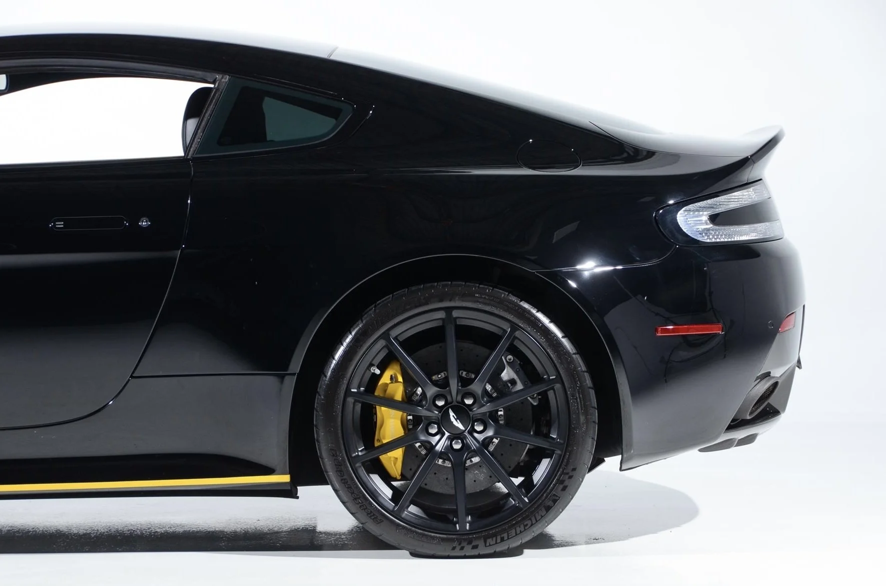 Close-up of the rear side of a black sports car, showing the tire, yellow brake caliper, and part of the rear light and aerodynamic design.