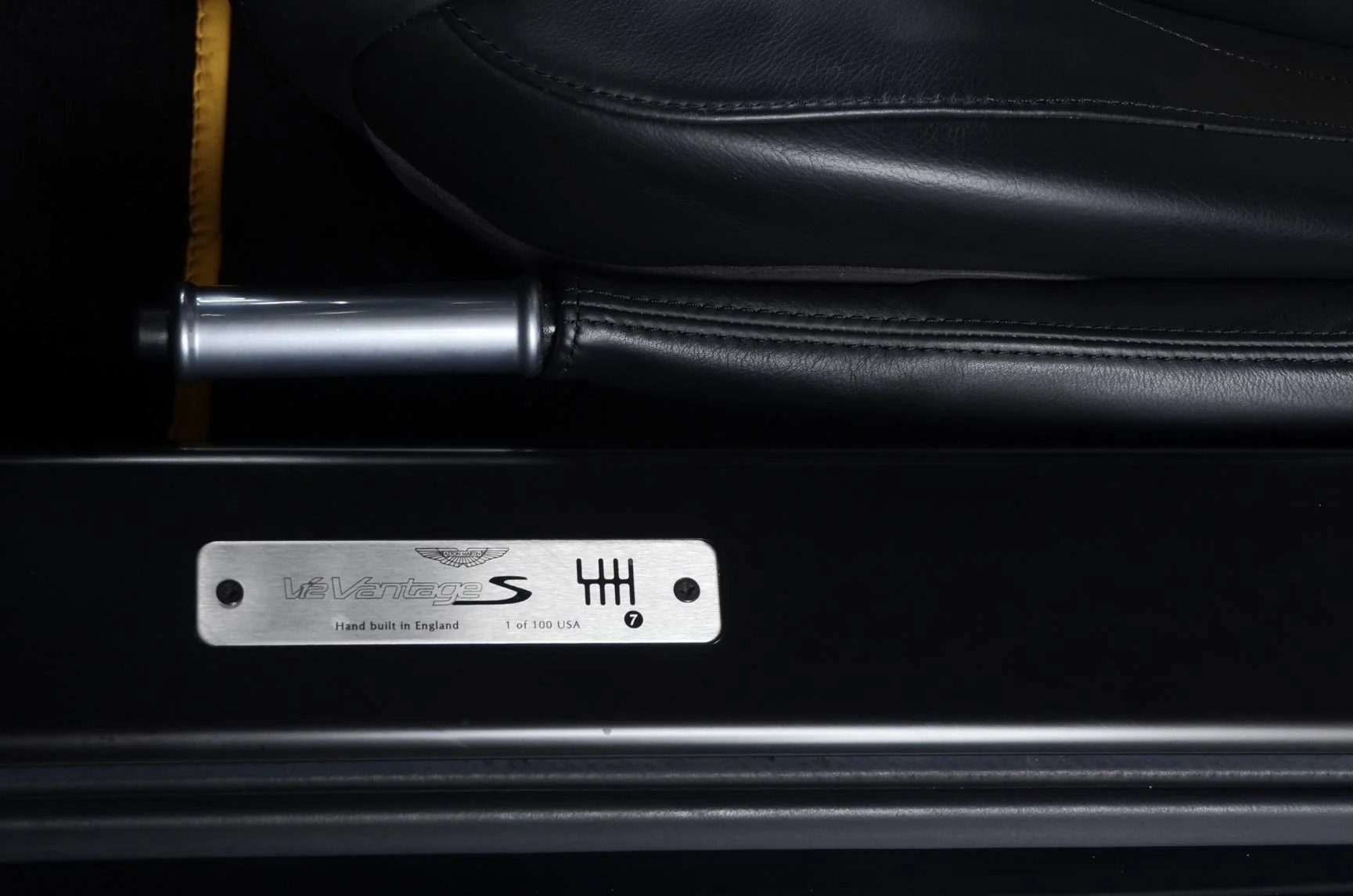 Close-up of a black leather Aston Martin V12 Vantage S car seat with a metal plaque that reads "V12 Vantage S. Hand built in England. 1 of 100 USA." The seat material has detailed stitching, and there is a metal bar visible at the front of the seat.
