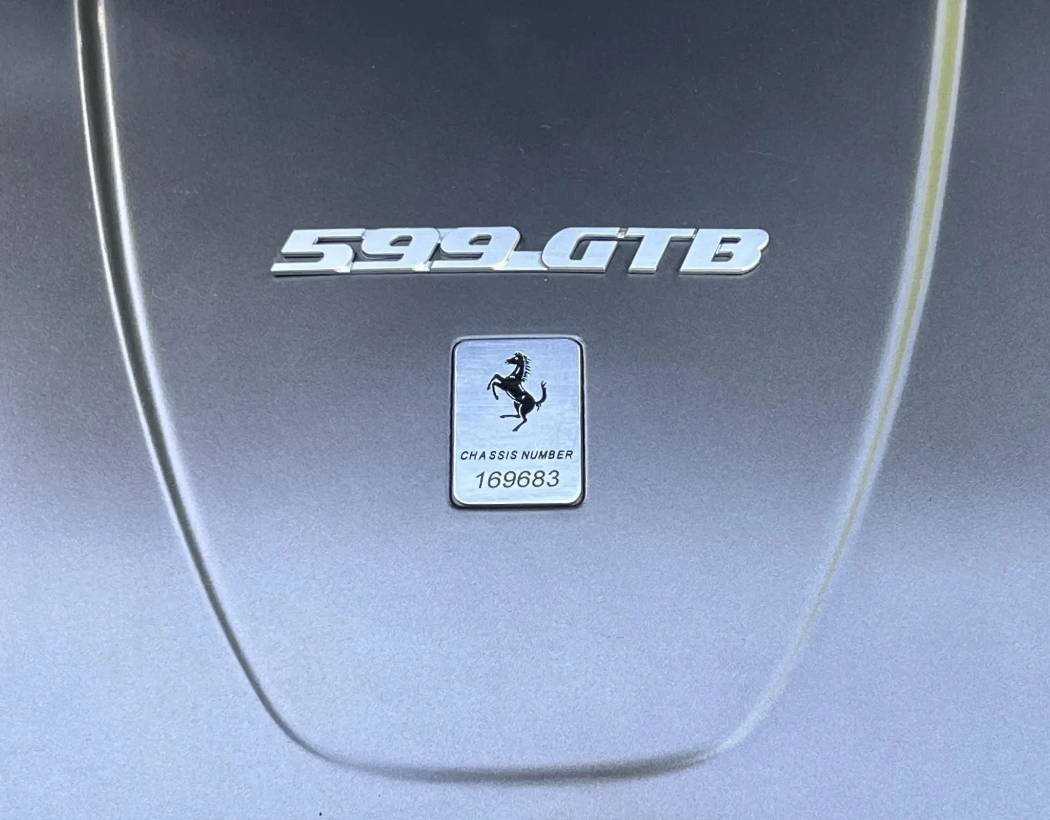Close-up of a silver Ferrari with a badge displaying '599 GTB' and a shield with a prancing horse logo and chassis number 169683.