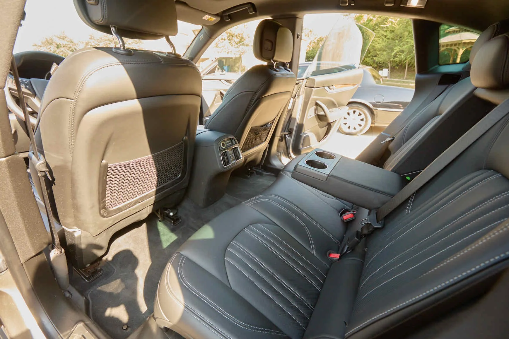 Interior of a vehicle showing the back seats, with black leather upholstery, seatbelts, and center cup holders, and the front seats with mesh storage on the back.