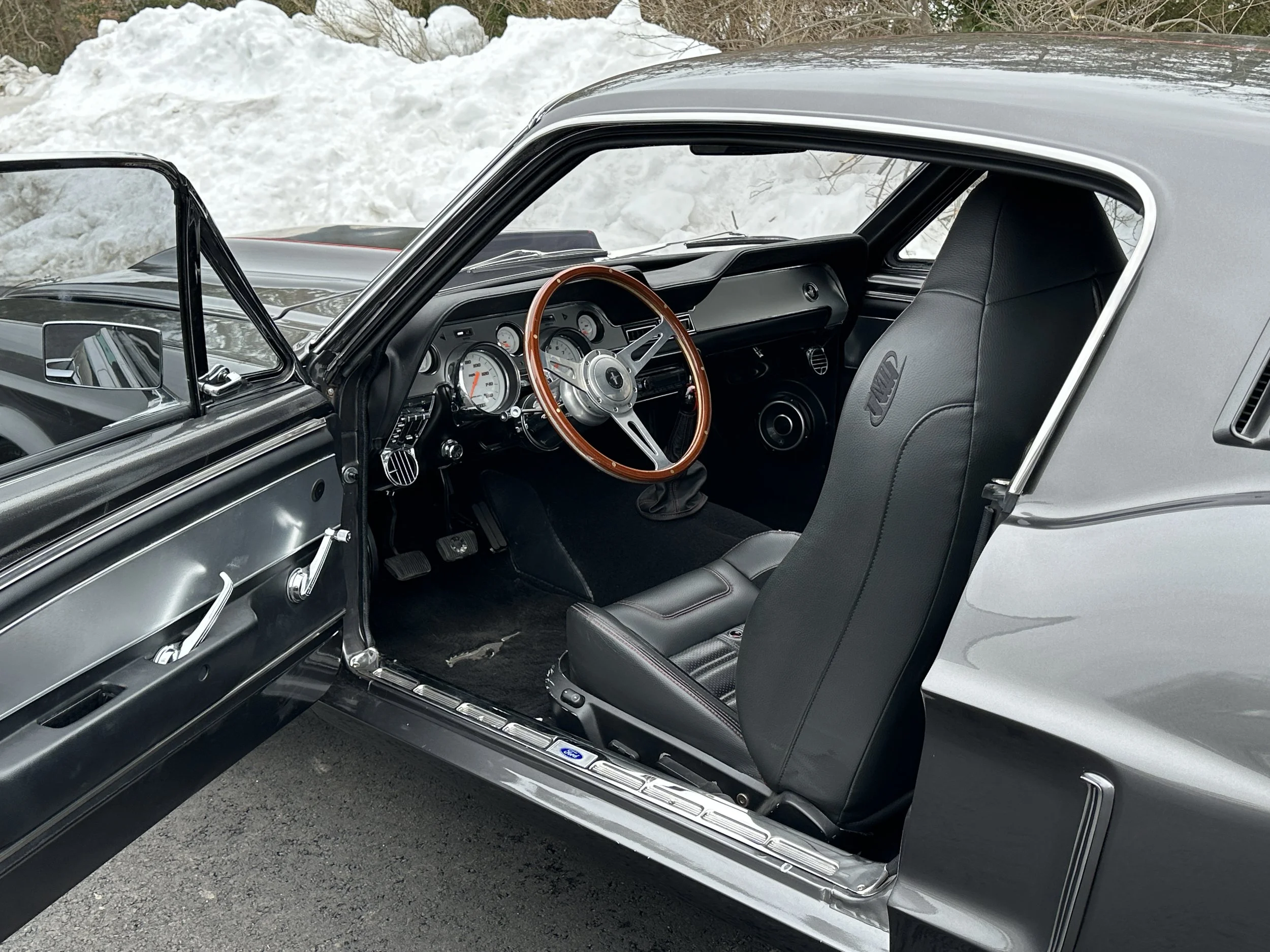 Interior of a vintage car with black leather seats and a wooden steering wheel, parked outside with snow in the background.