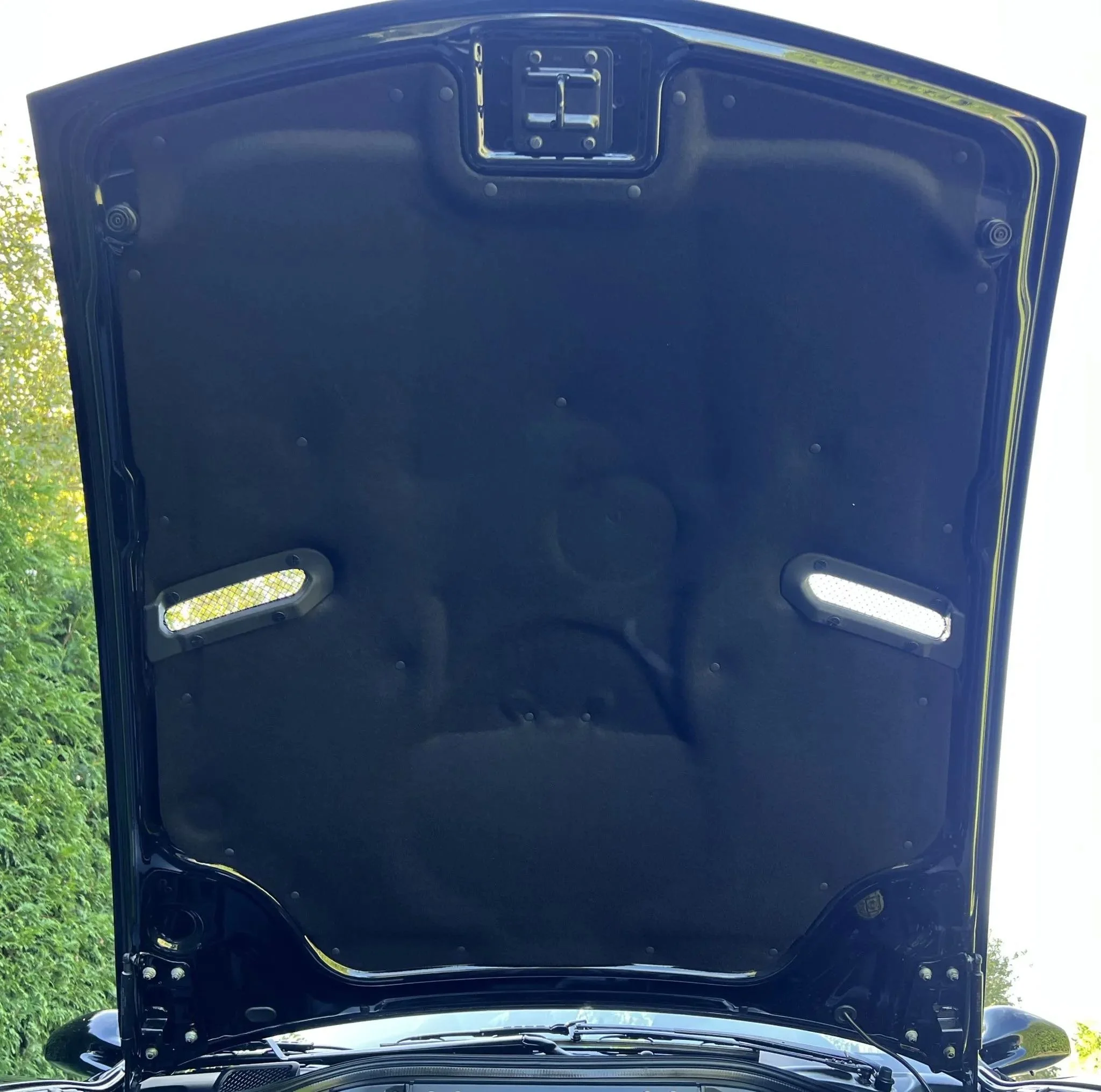 The underside of a car's hood, which is black with some reflective surfaces, showing the hood latch at the top center and two vents on either side.