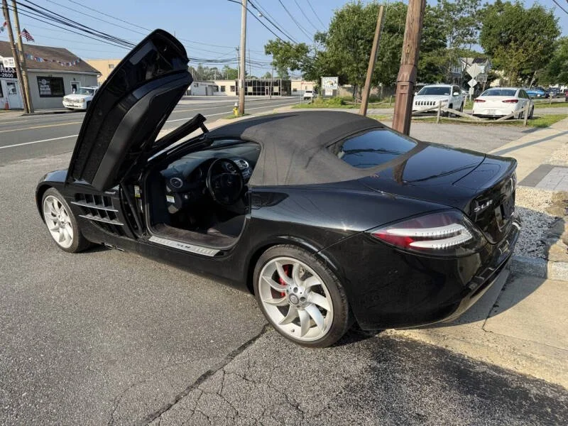 Black convertible sports car with gullwing door open, parked on street near sidewalk with other cars and trees in the background.