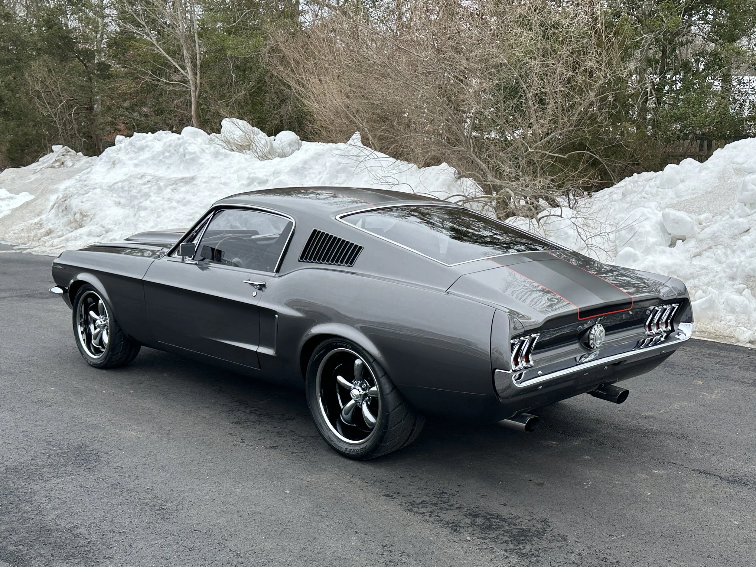 A classic gray muscle car parked on a paved road with snow piled along the side and trees in the background.