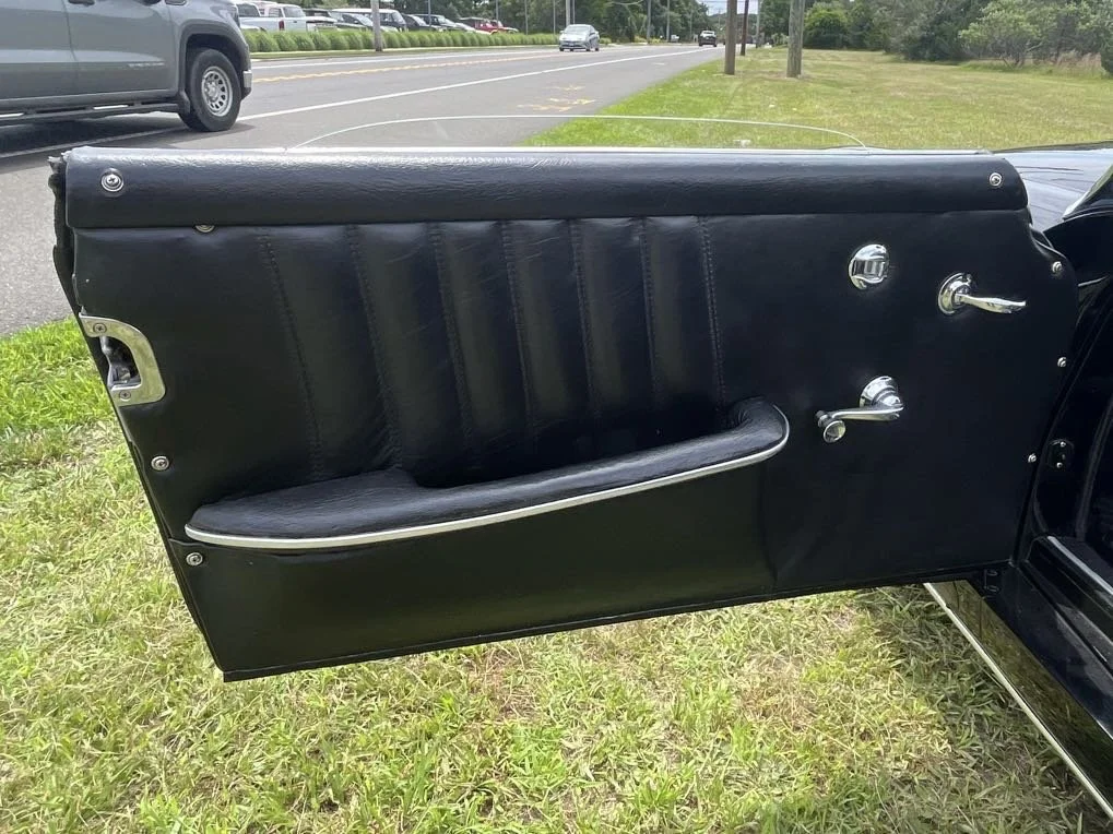 Close-up of a vintage car door with black upholstery, window crank, door handle, and armrest, parked outdoors on grass.