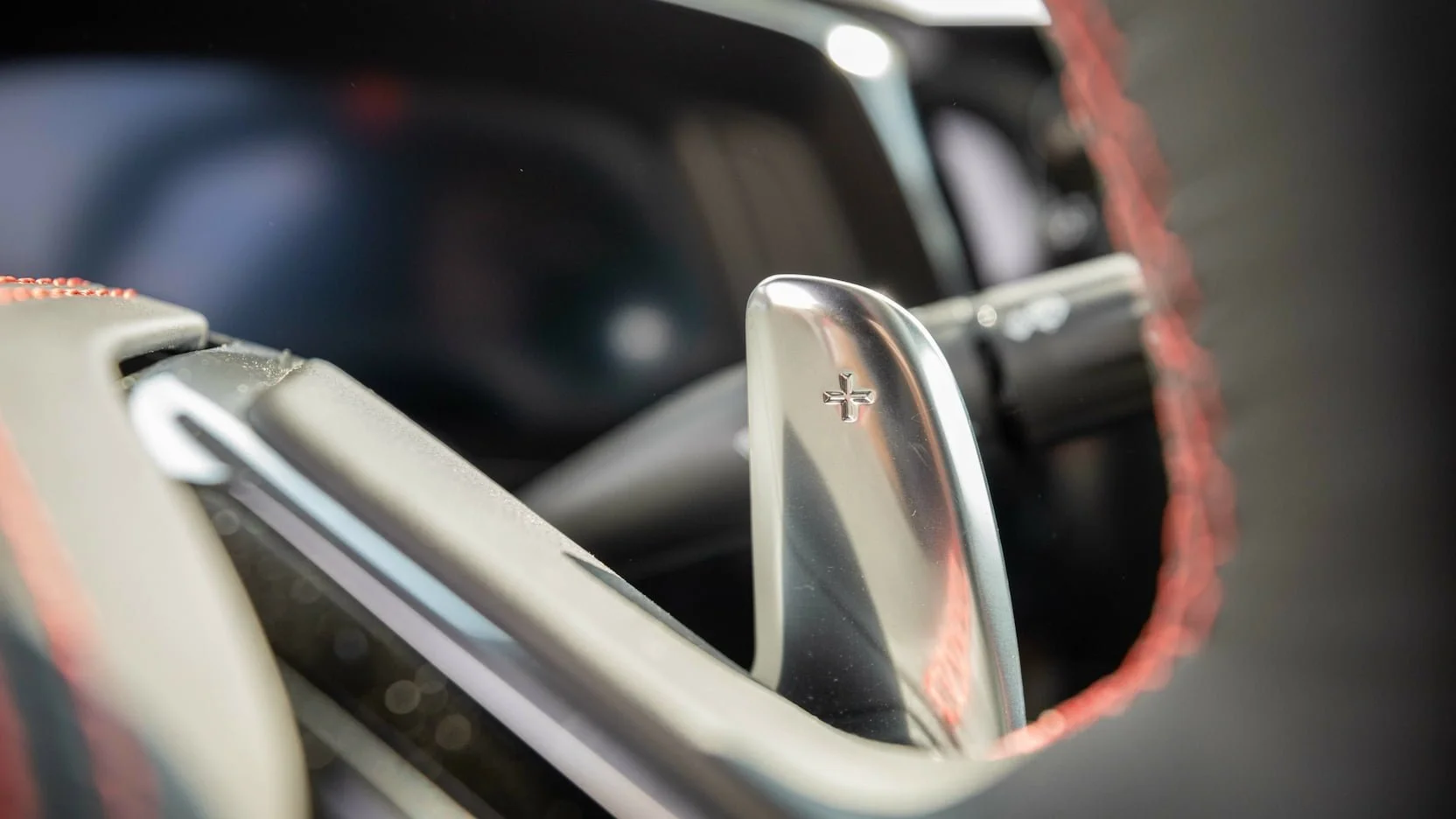 Close-up of a paddle shifter in a car, with a plus (+) sign visible, indicating an upshift, with a red stich steering wheel in the background.