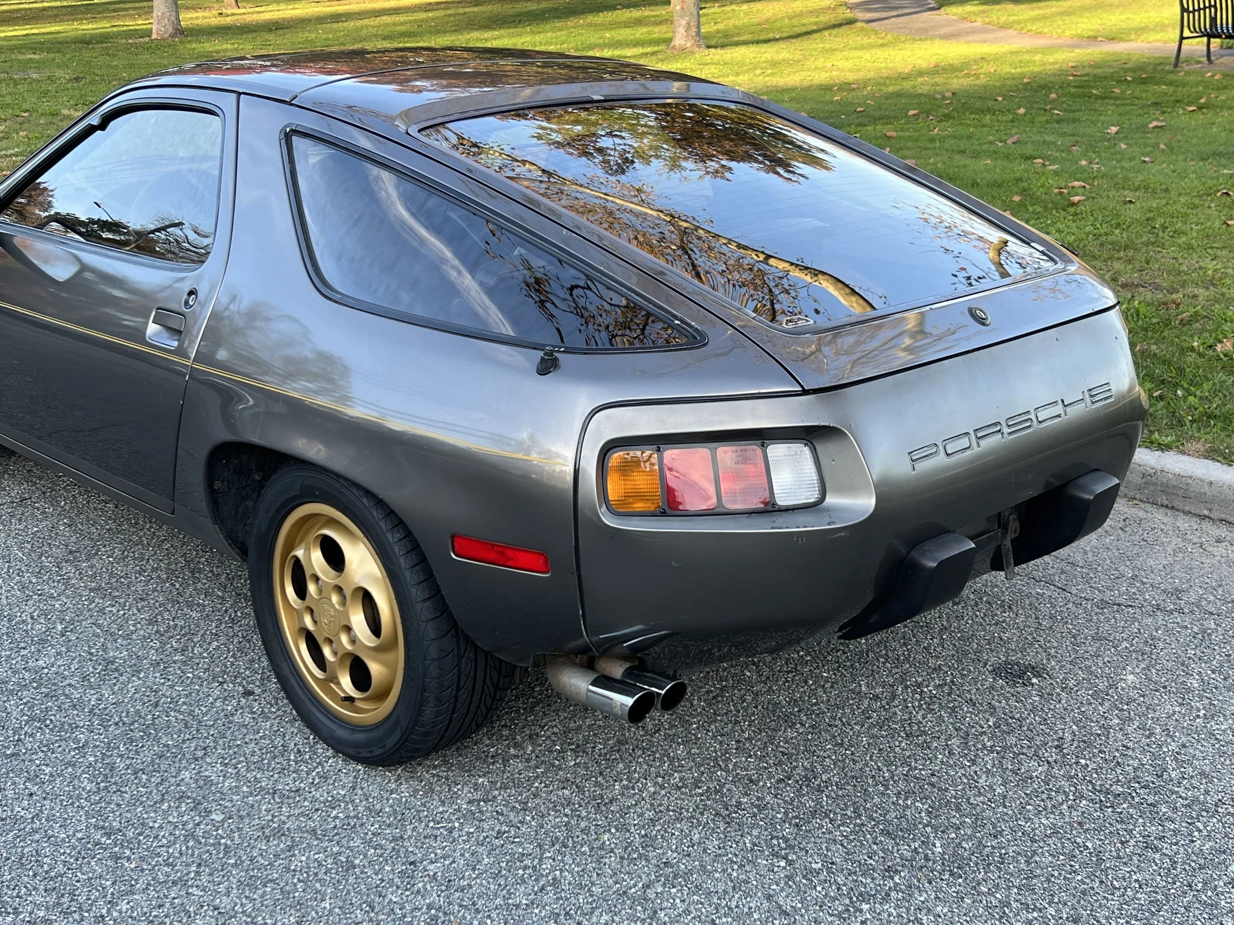 A classic gray Porsche 928 sports car parked on a paved surface with a grassy park area and trees in the background, featuring gold wheels and dual exhaust pipes.