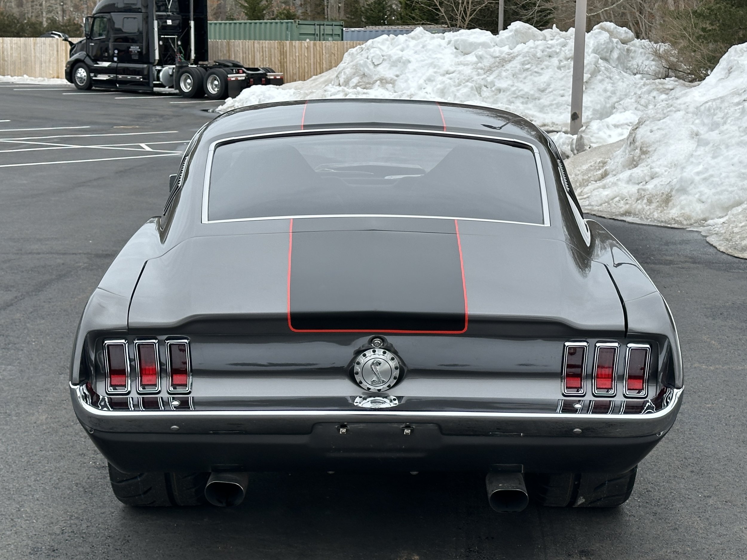 Rear view of a classic black muscle car with red racing stripes, parked in a parking lot with snow piles in the background.