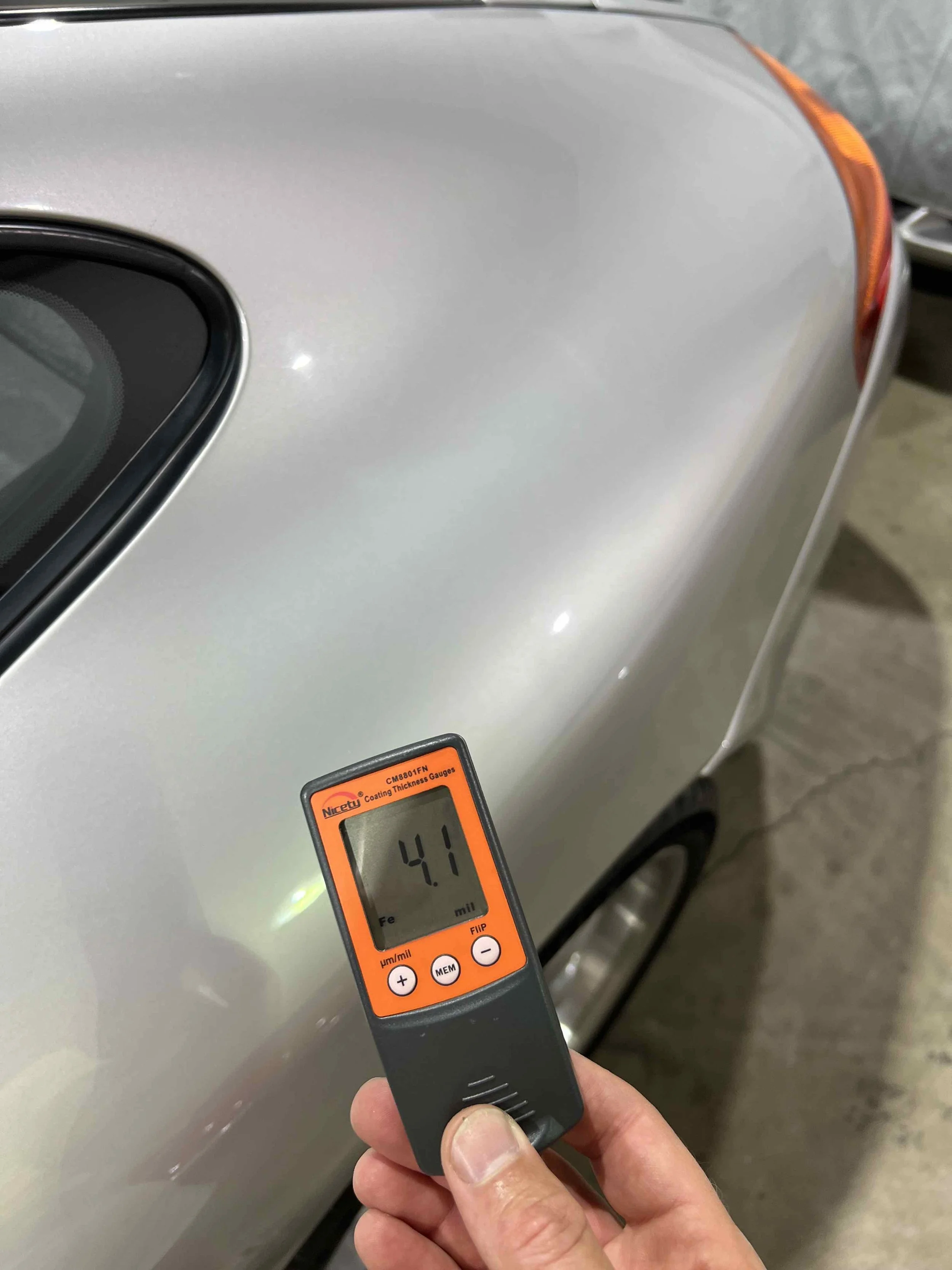A person holding a coating thickness gauge reading 4.1 micrometers against the side of a silver car in a garage.